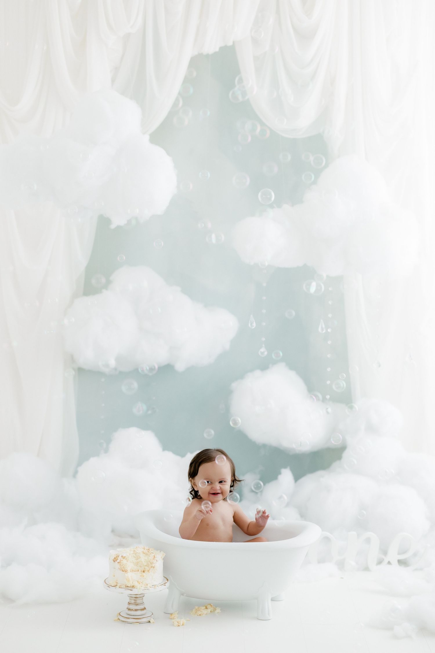Baby smiling in a white bathtub surrounded by fluffy clouds, bubbles, and white curtains in a dreamy studio setup.