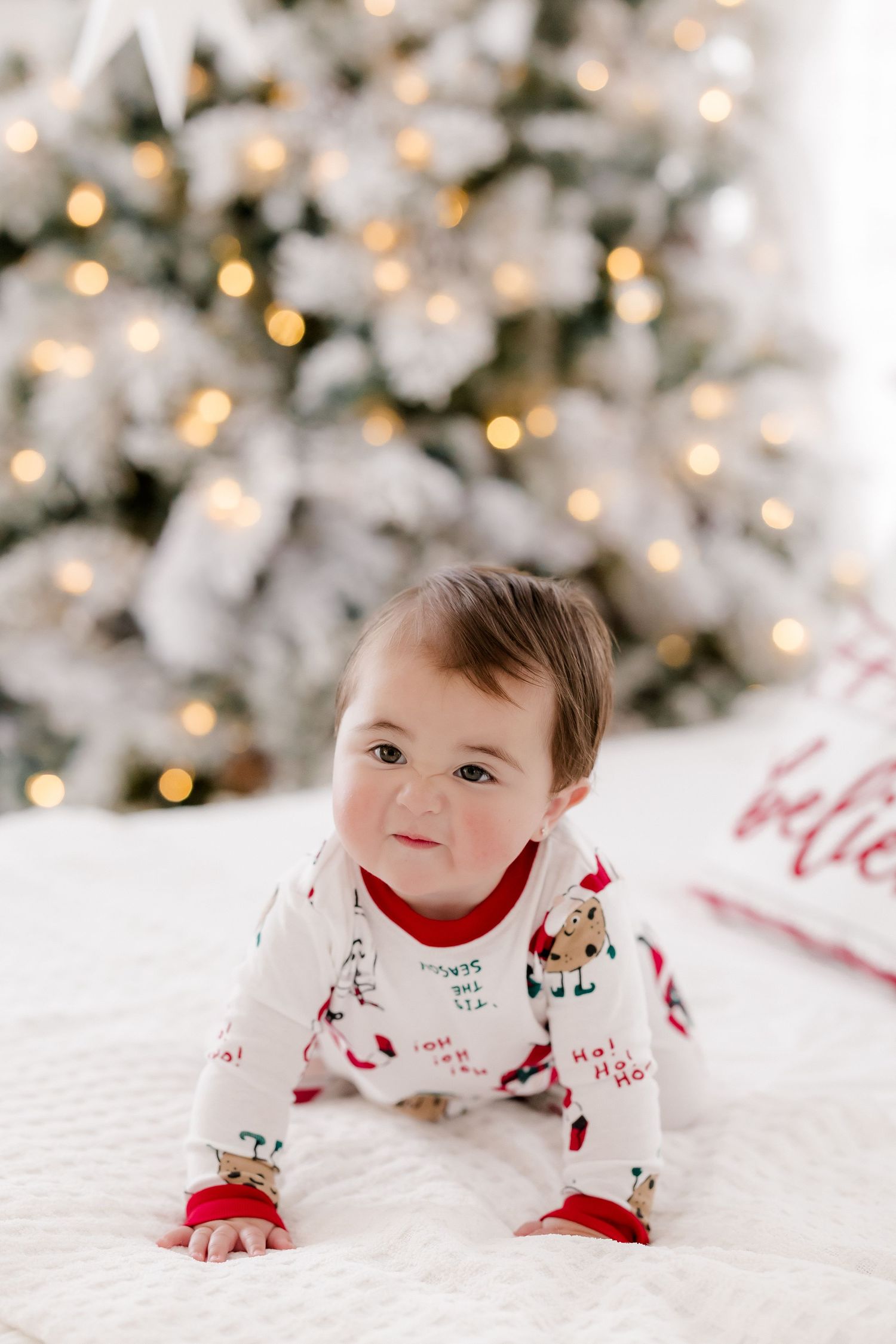 Baby in Christmas pajamas crawling on white blanket in front of lit flocked holiday tree.