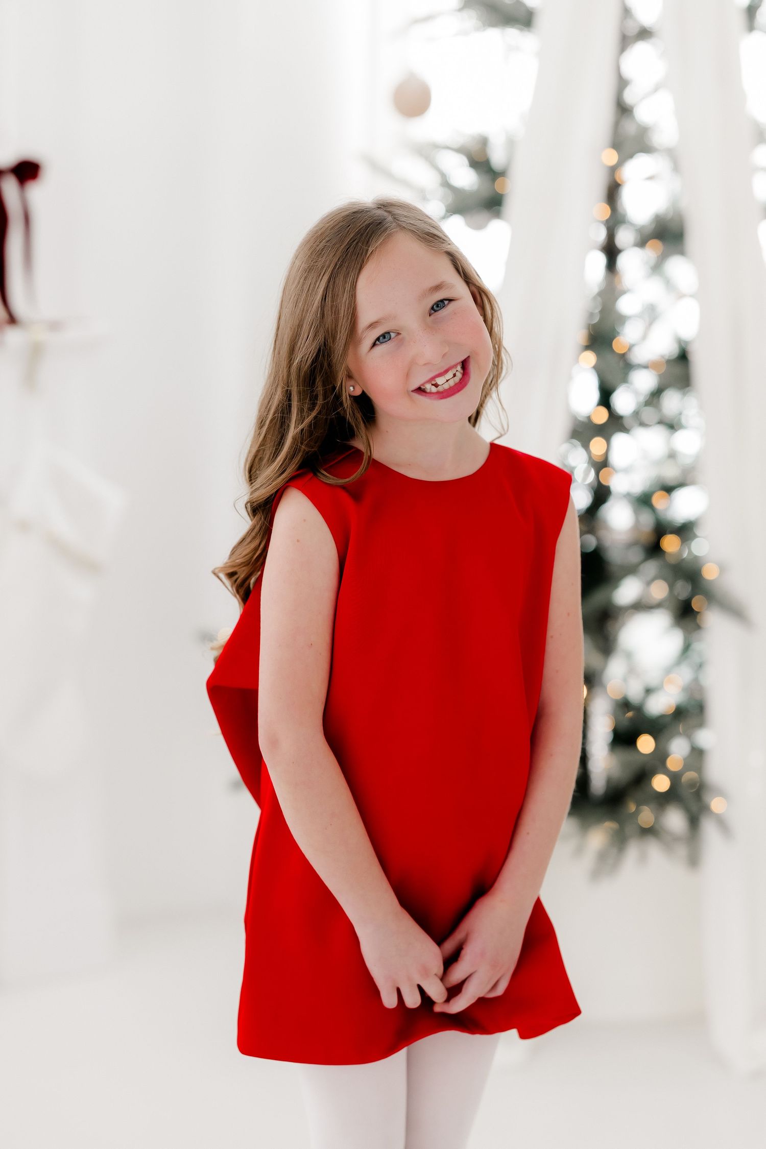 Young girl in red dress smiling joyfully in front of a decorated Christmas tree in a white room.