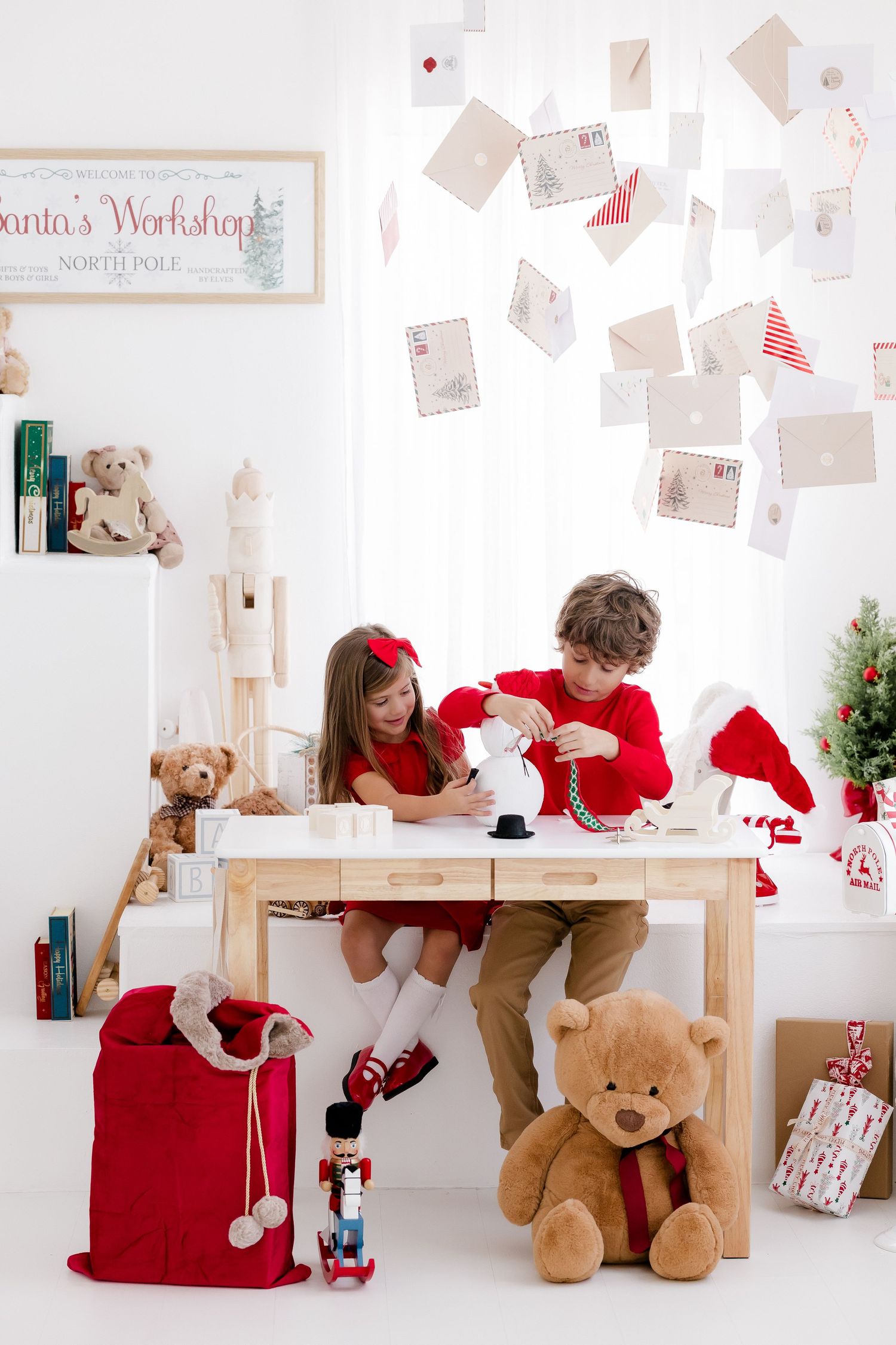 Two children in red outfits play at a wooden table in a festive Santa's Workshop setting with teddy bears and Christmas decor.