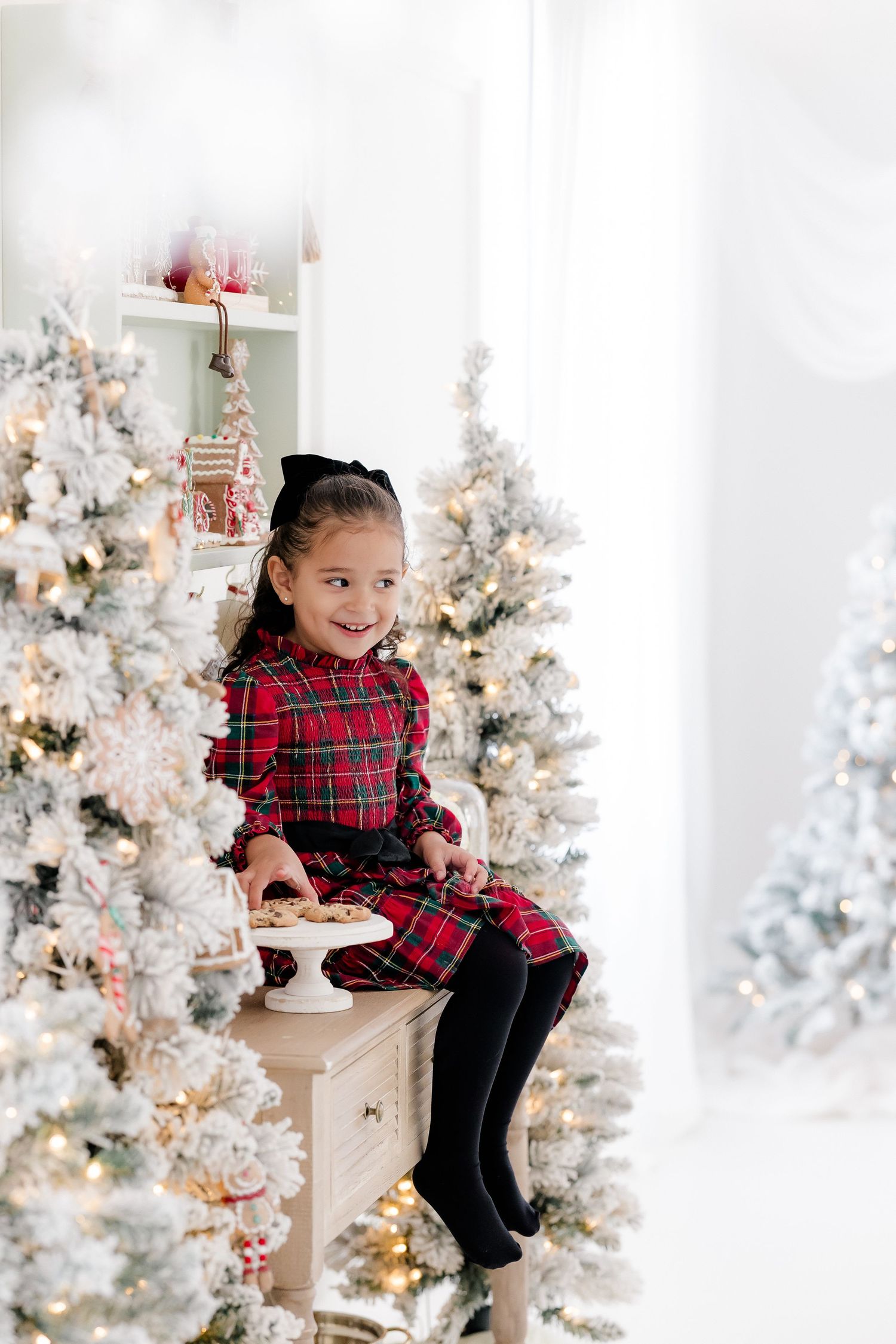 Young girl in red plaid dress sitting among flocked Christmas trees with white lights in festive holiday setting.