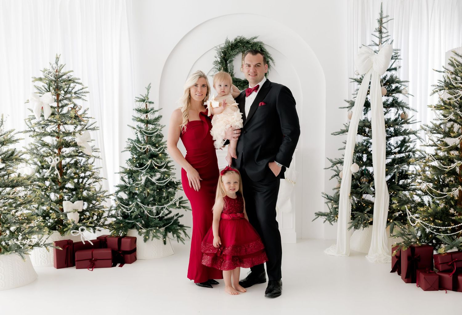 Family of four in red and black formal wear posing among snow-flocked Christmas trees with wrapped gifts.
