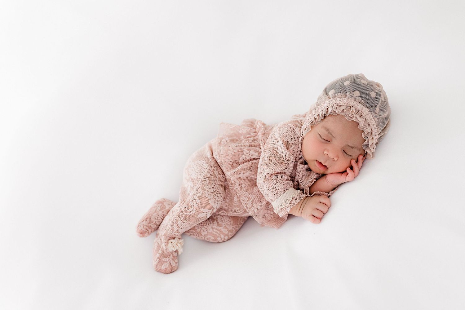 Sleeping baby wearing pink lace outfit and bonnet during professional photoshoot.