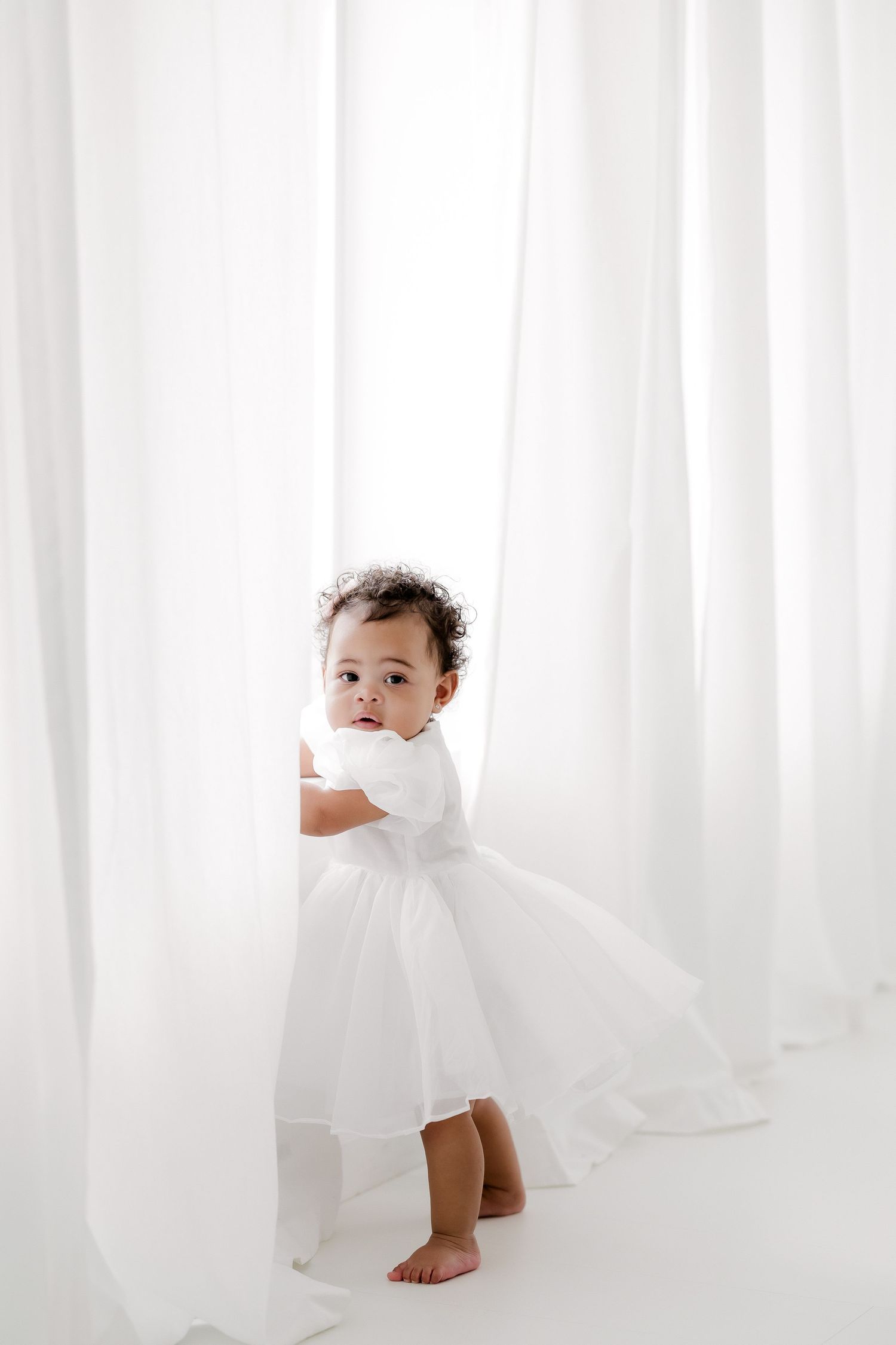 A small child in a white outfit plays with flowing sheer curtains in a bright minimalist setting.