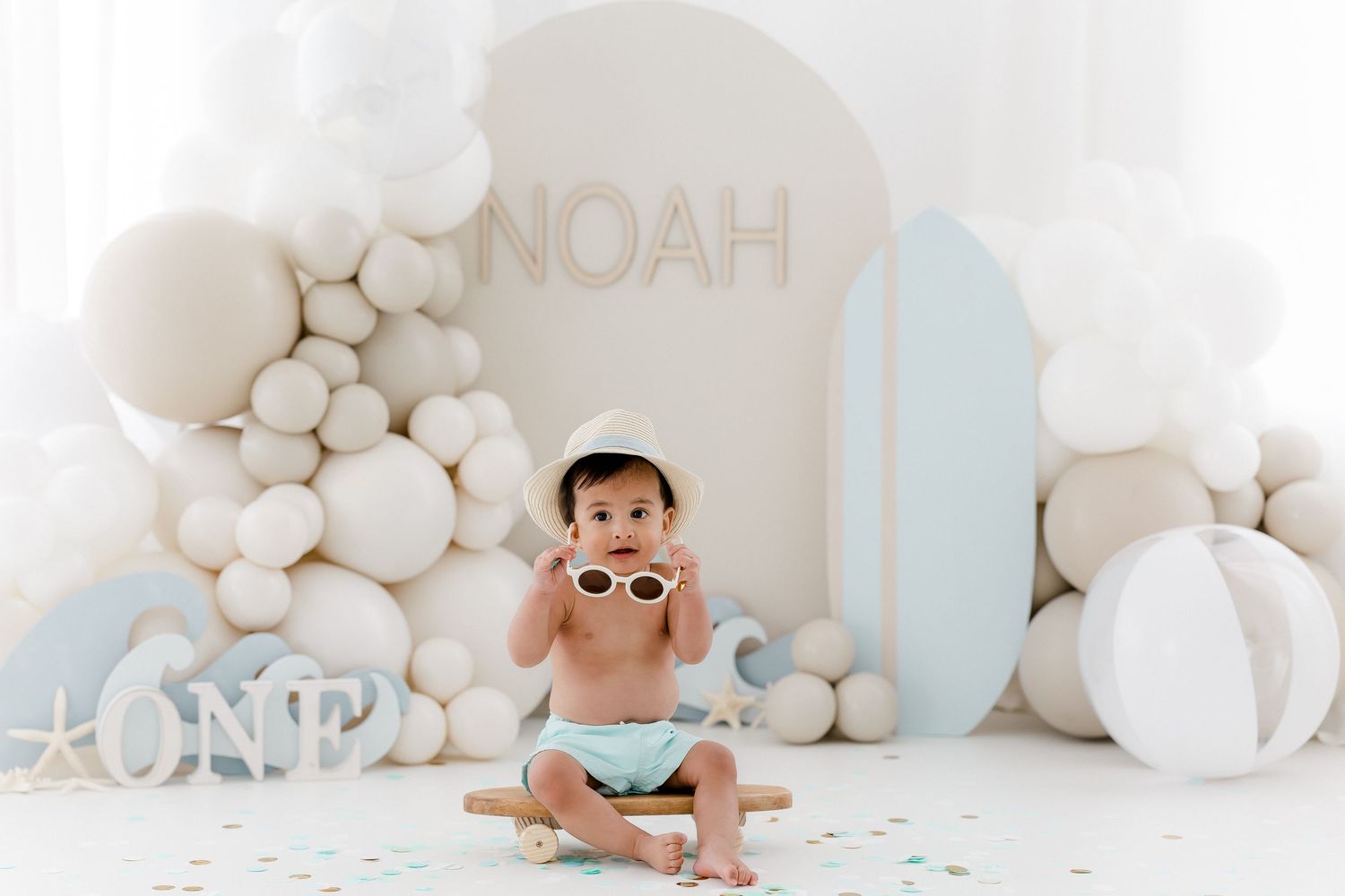Baby in white diaper crawls across white balloon-decorated backdrop during professional photo session.