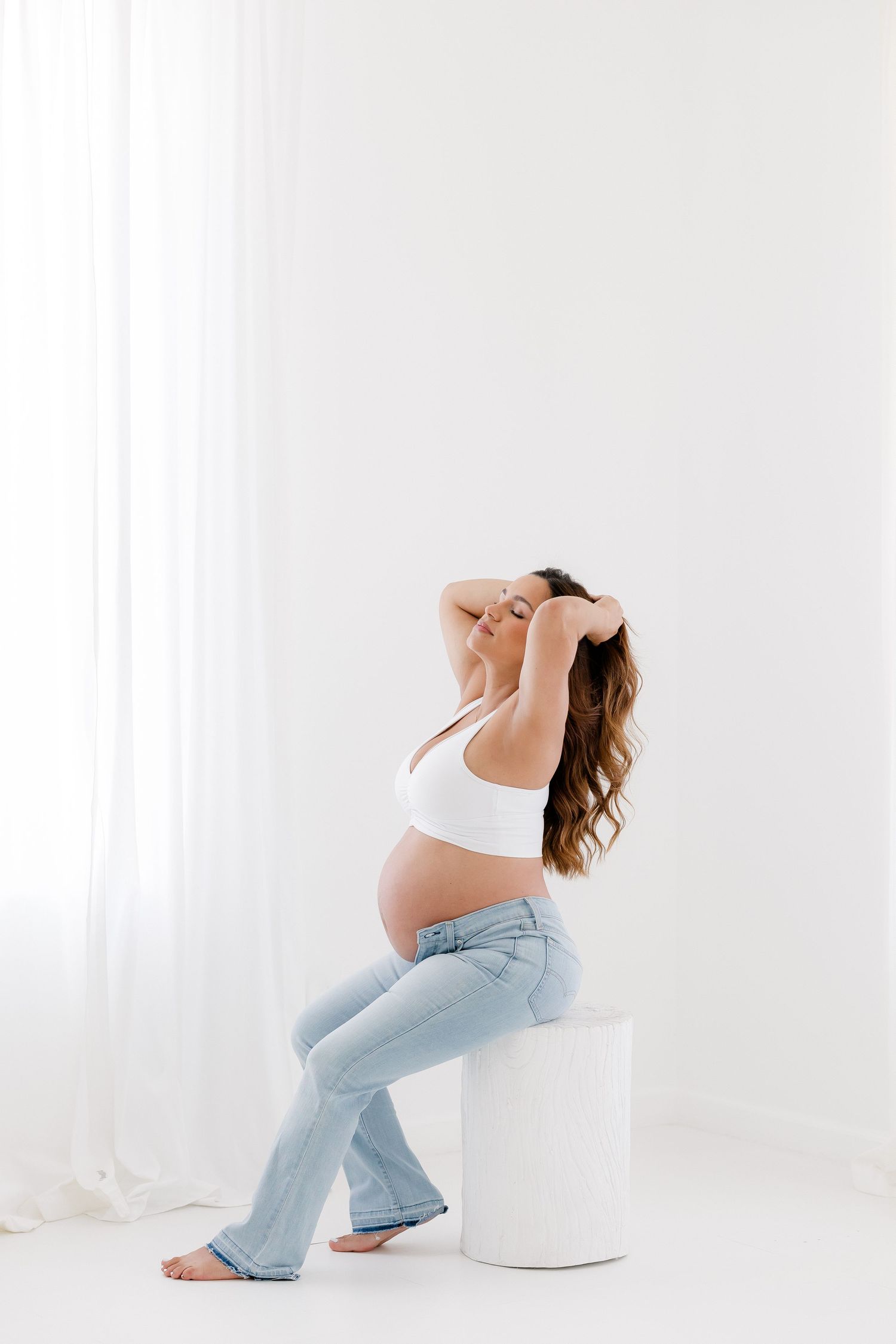 Pregnant woman in white sports bra and light blue jeans poses gracefully in minimalist white studio setting.