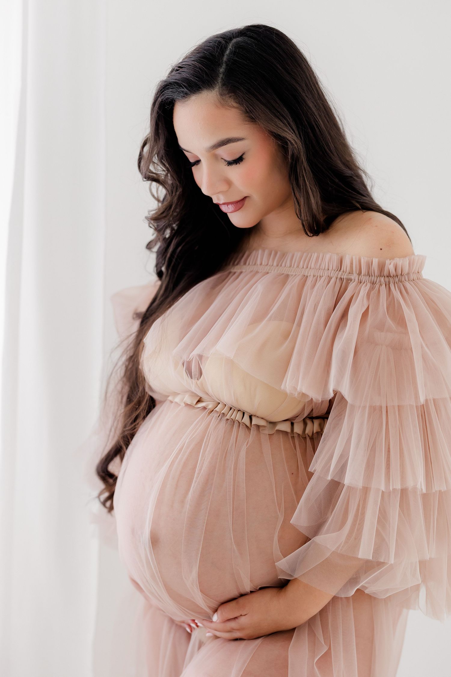 Expectant mother in flowing blush pink tulle off-shoulder maternity gown poses against white backdrop.