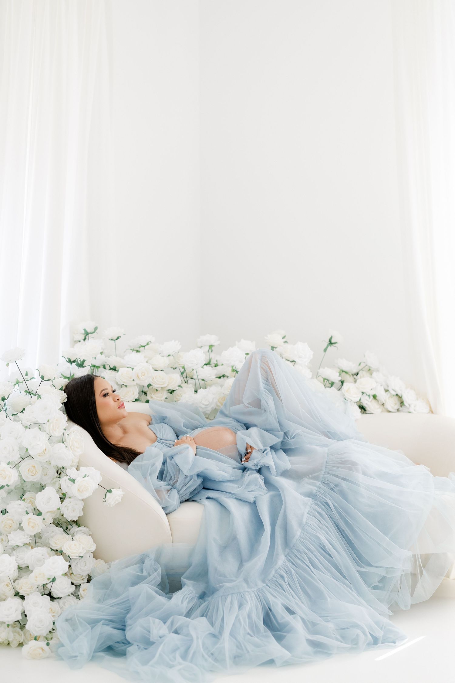 Dreamy maternity portrait with white roses and baby blue gown in studio setting.