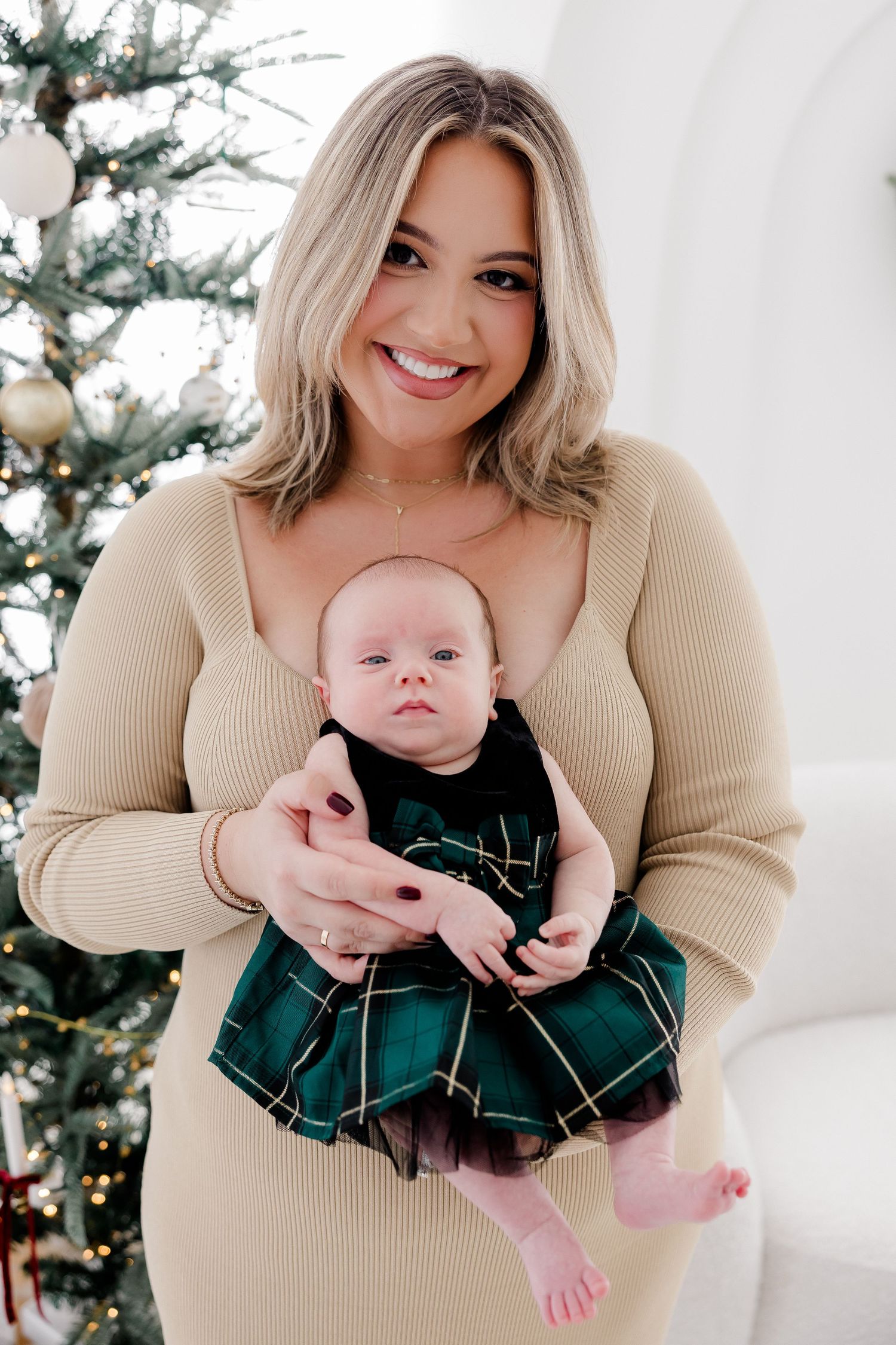 Mother and baby in matching cream outfits pose for holiday photos with decorated Christmas tree.
