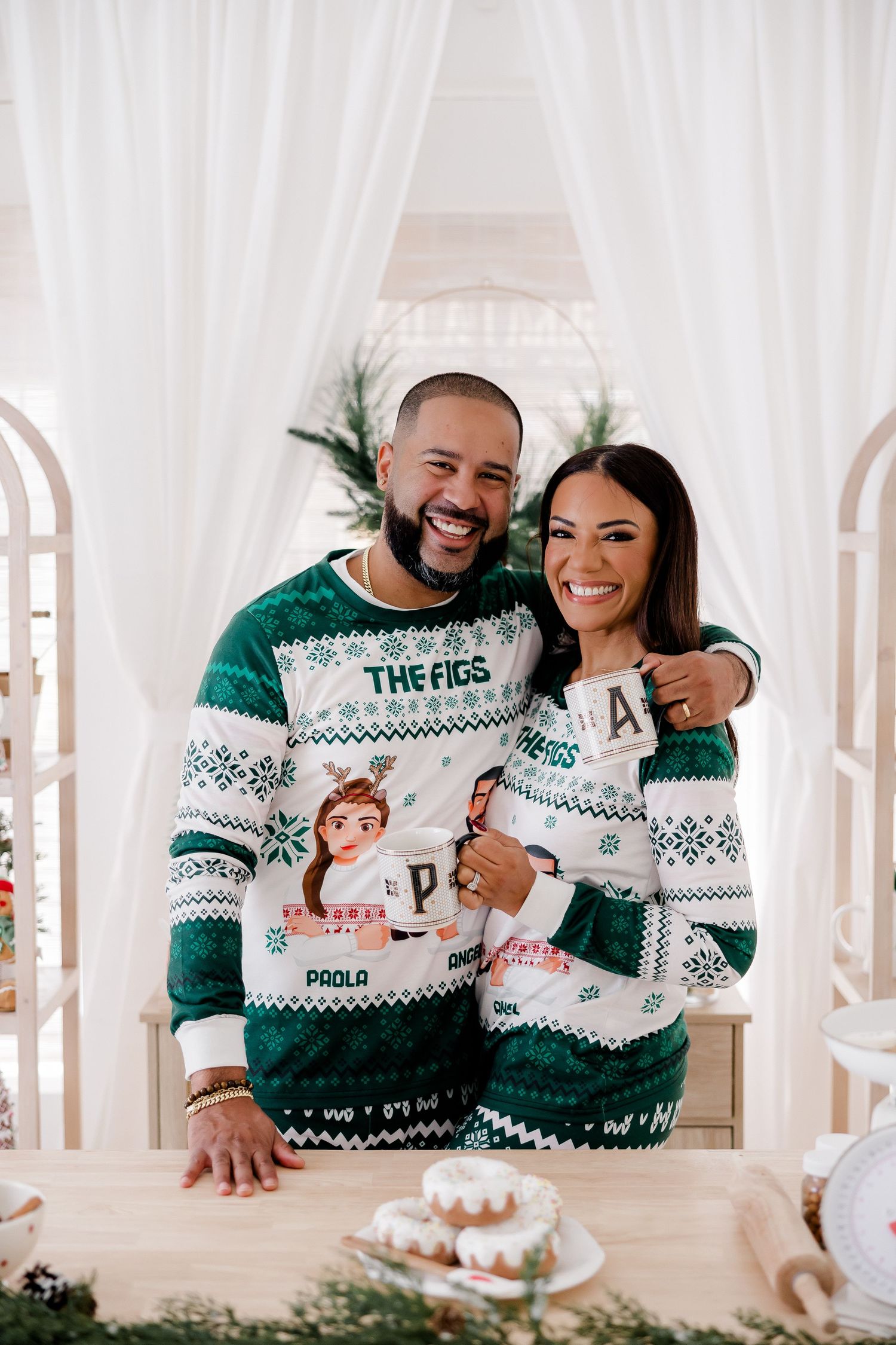 Couple wearing matching green and white Christmas sweaters smile together in front of white curtains.