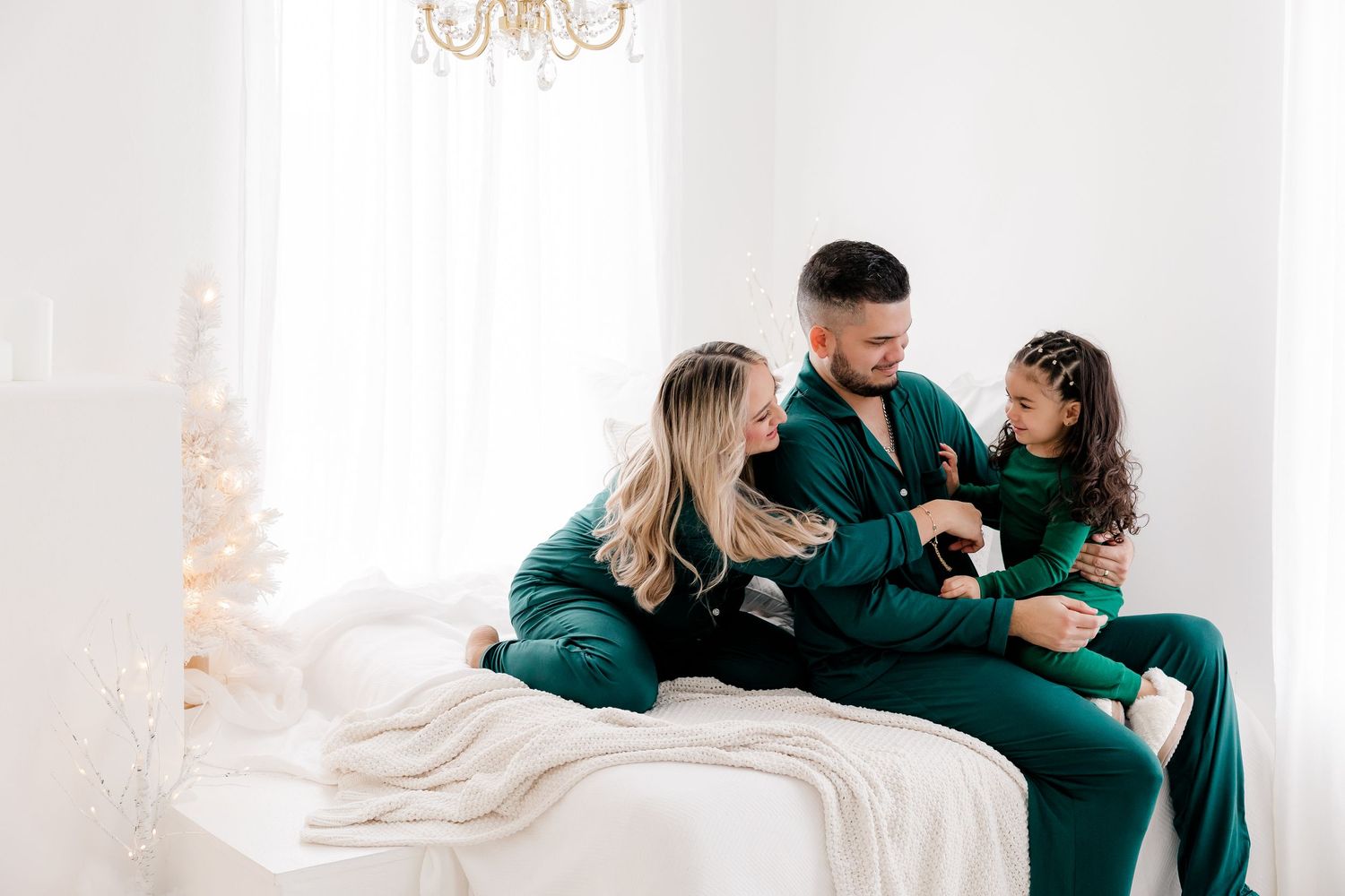 Family in matching emerald outfits sharing intimate moment on white bed with Christmas decor.