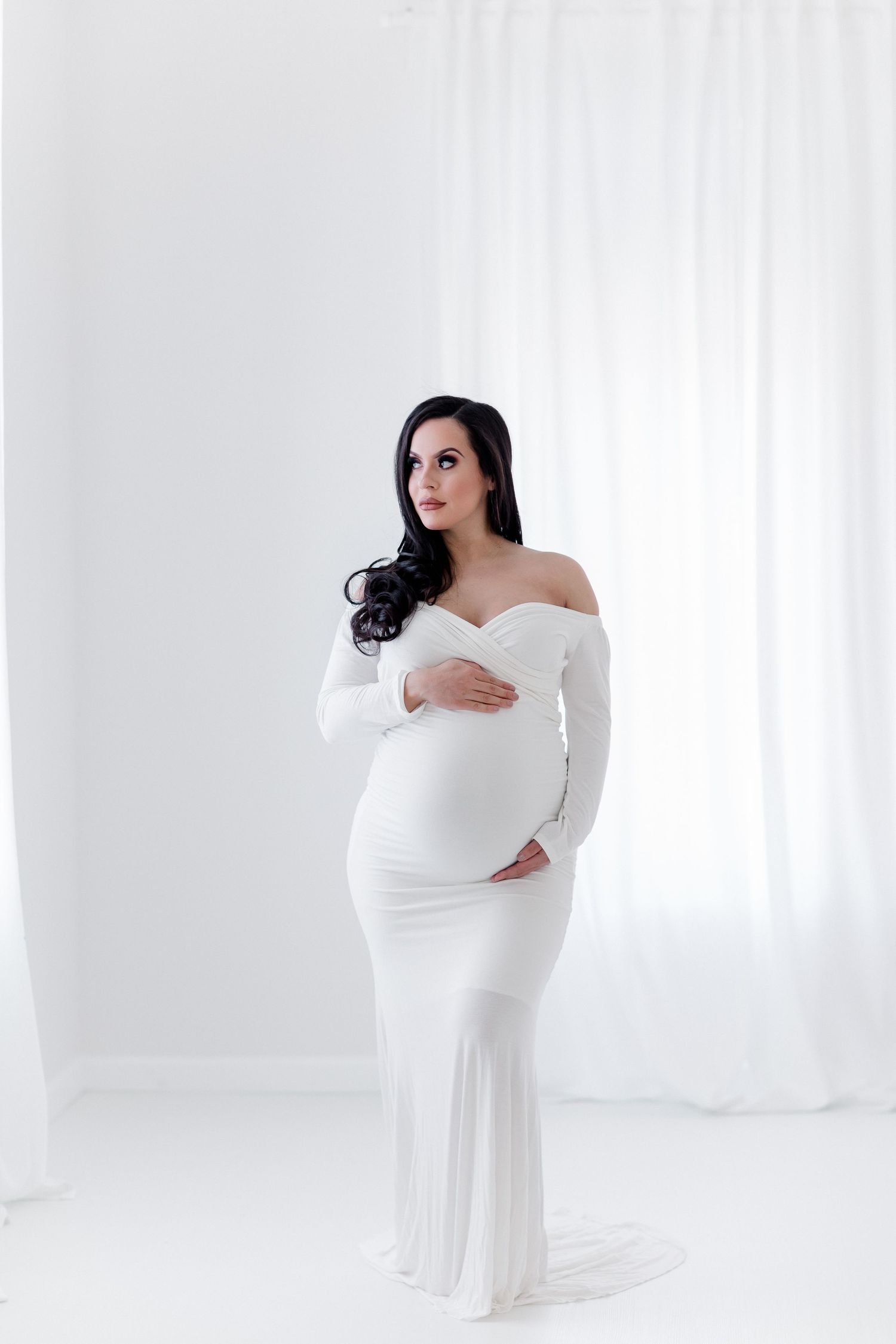 Classic maternity portrait in fitted white off-shoulder gown against bright white backdrop.