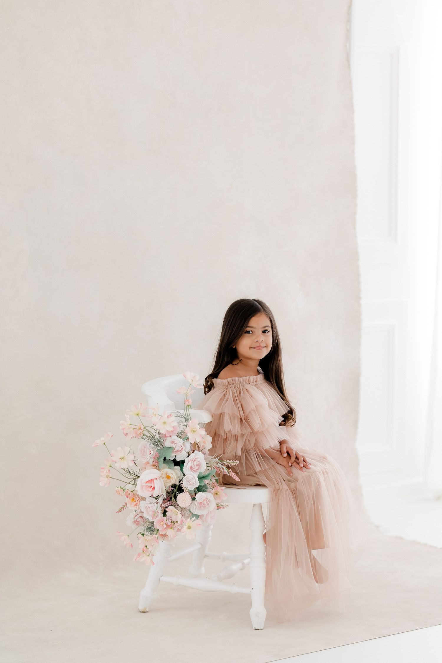 A child in a flowing blush pink dress sits elegantly on a white chair with a pastel floral bouquet.