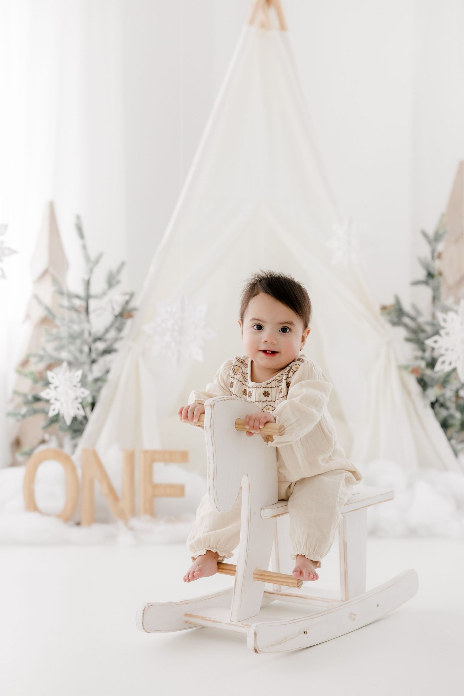 Baby in white outfit sits in minimalist winter wonderland setup with teepees and silver decorative trees.