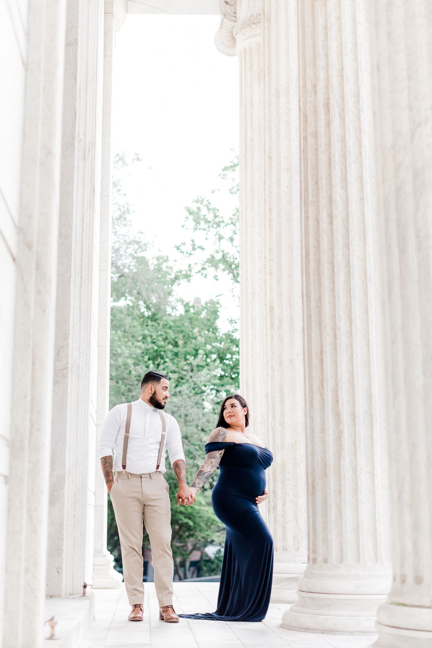 Pregnant woman in navy dress poses elegantly between white classical columns during maternity photoshoot.
