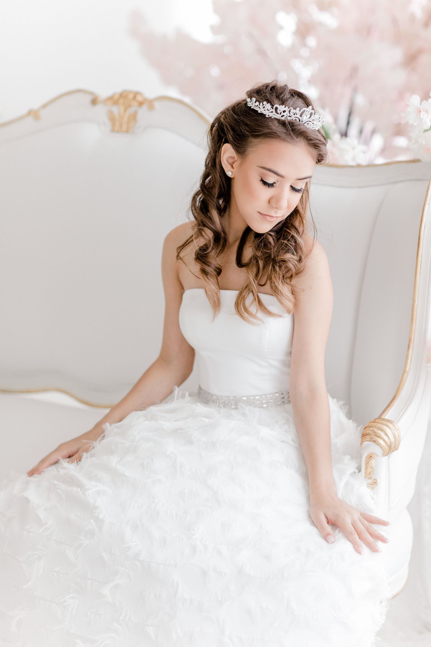 A bride in a white strapless ballgown and tiara sits elegantly on an ornate vintage chair with blush floral backdrop.