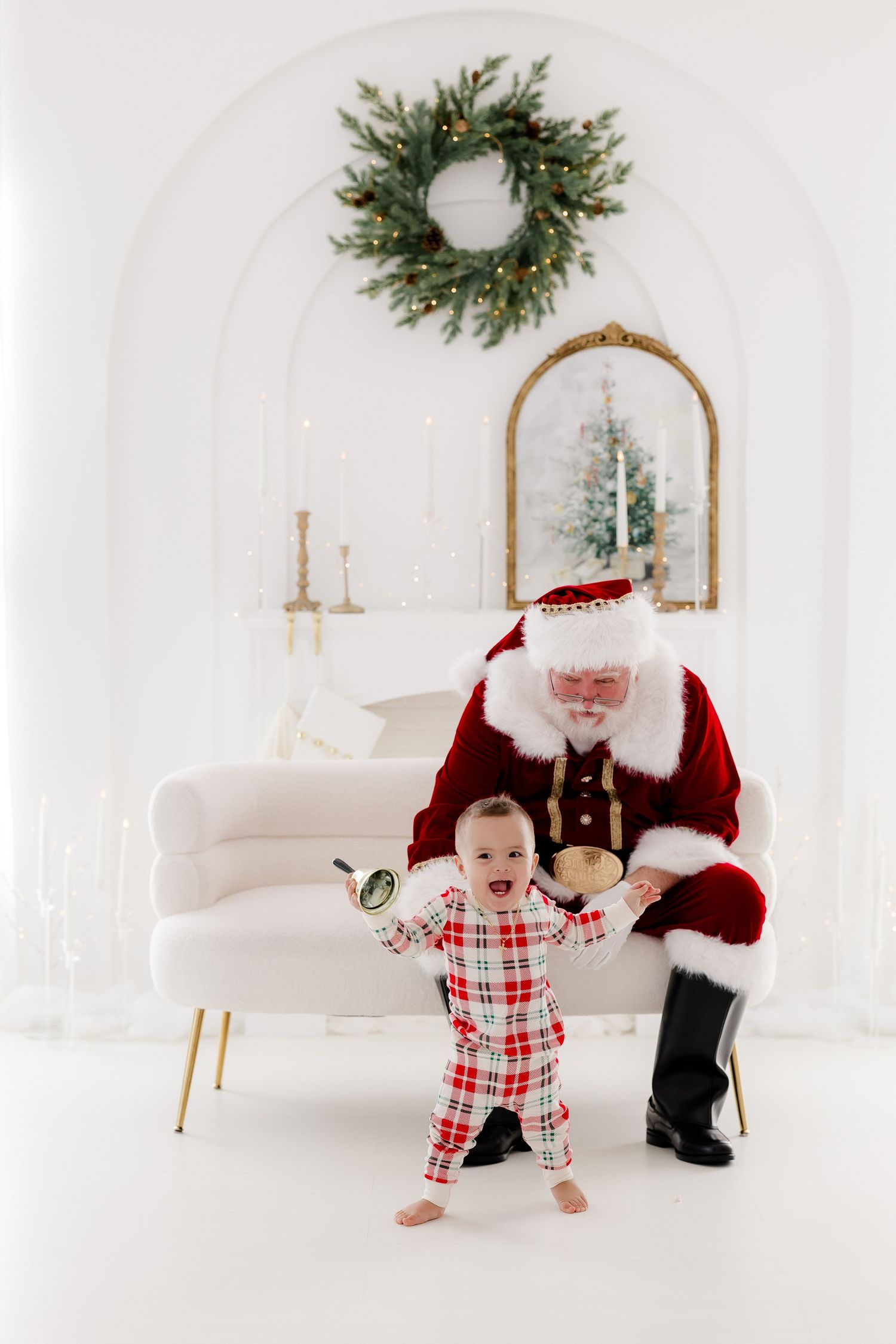 Joyful baby in plaid pajamas stands with Santa Claus on white sofa, Christmas wreath decorating festive background.