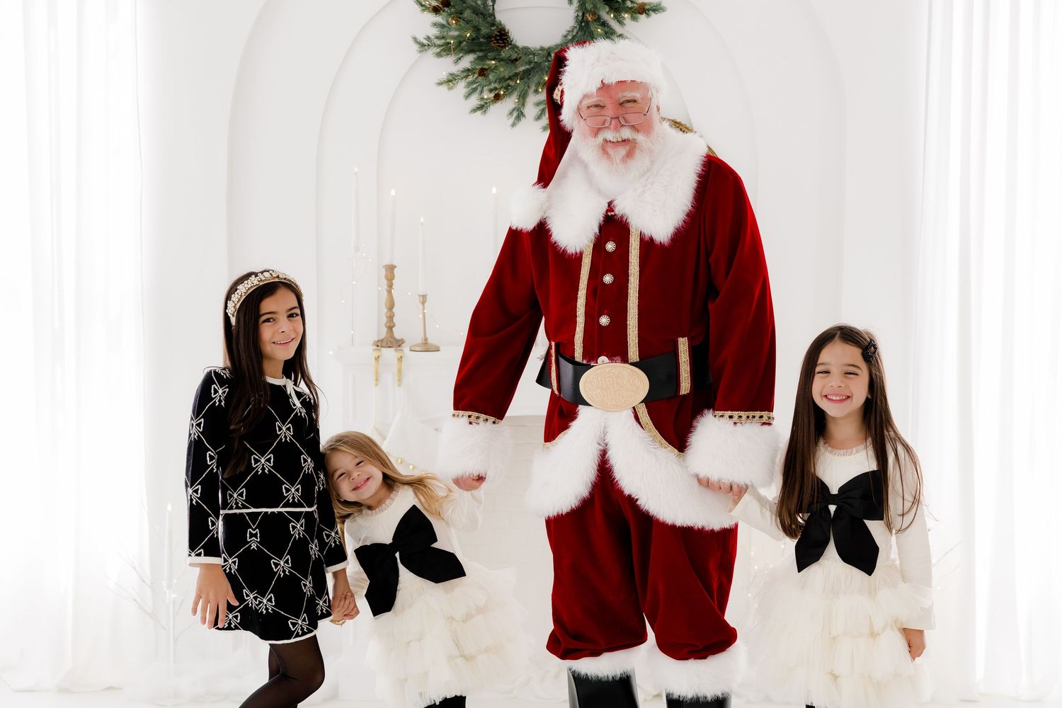 Three girls pose with Santa Claus in festive outfits against a white backdrop with holiday wreath decor.