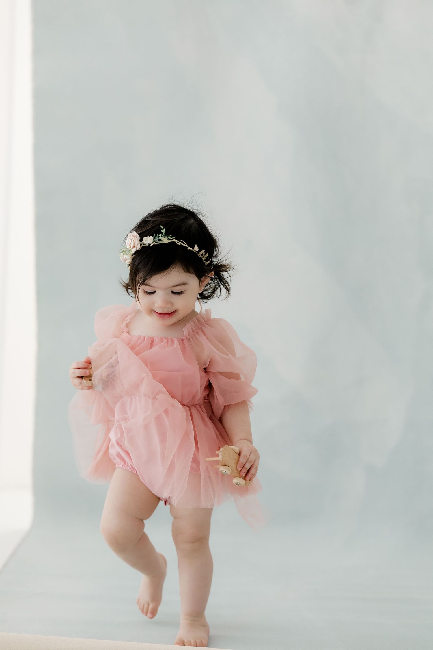A baby wearing a pink tulle dress plays and moves around during a studio photoshoot against a light gray background.