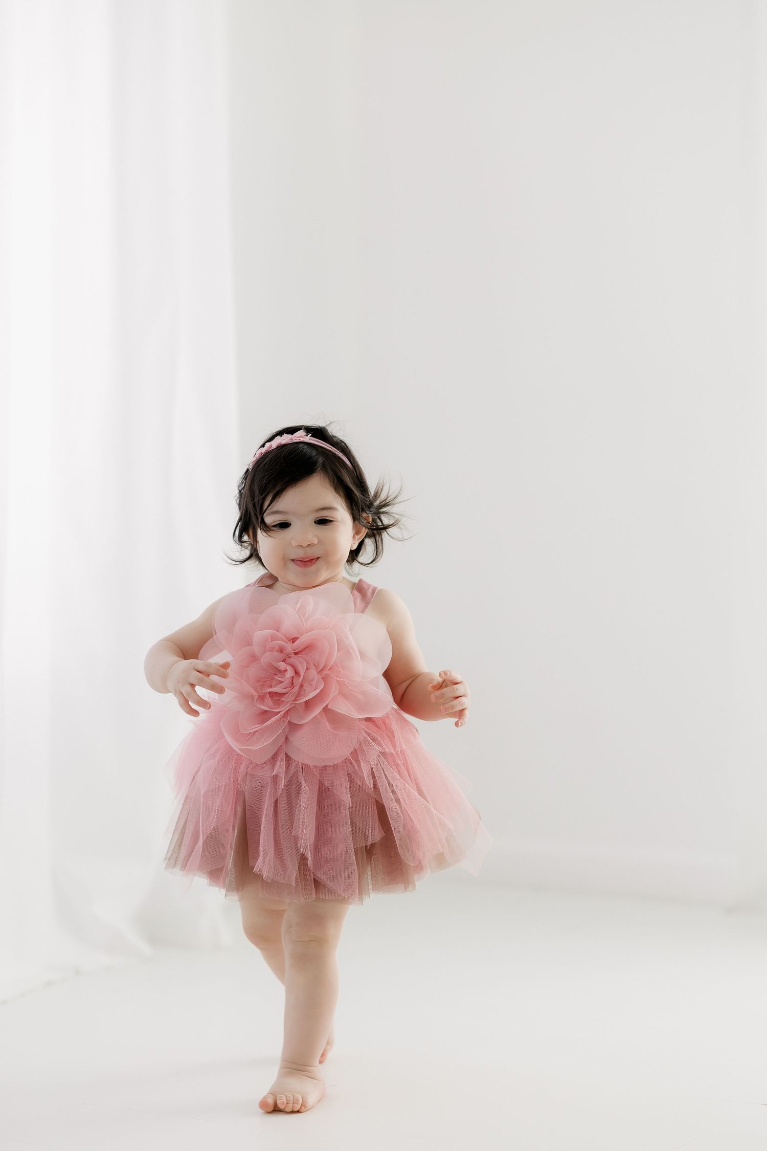 A small child in a pink tulle dress with a large floral detail dances barefoot in a bright white studio setting.