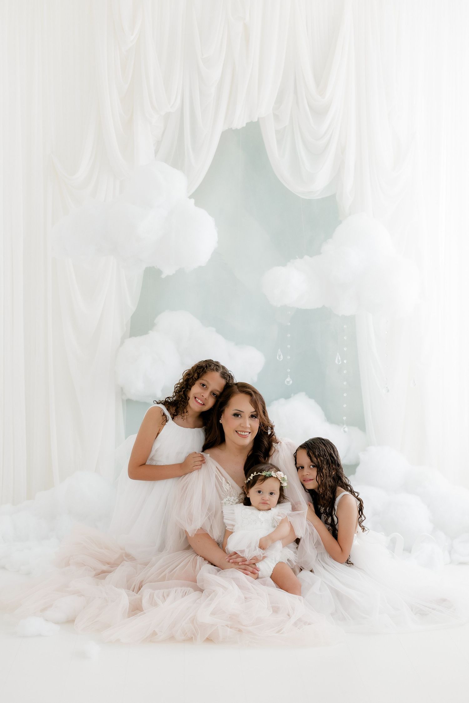 A mother and three daughters in white dresses pose together in a dreamy studio with cloud decorations and soft draping.