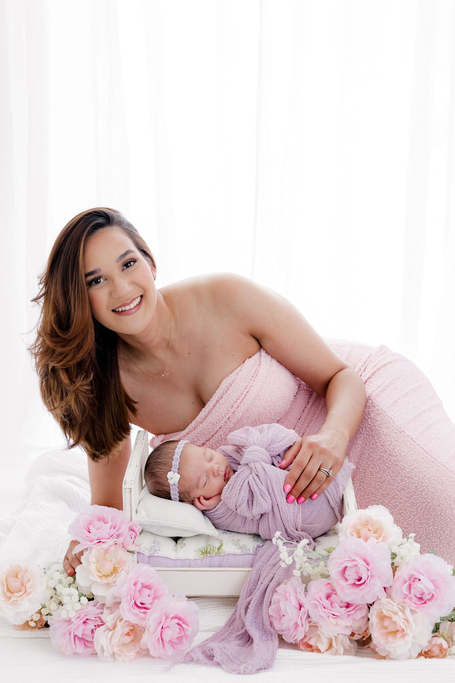 Elegant newborn photography with pink roses and soft mauve fabrics in bright studio setting.