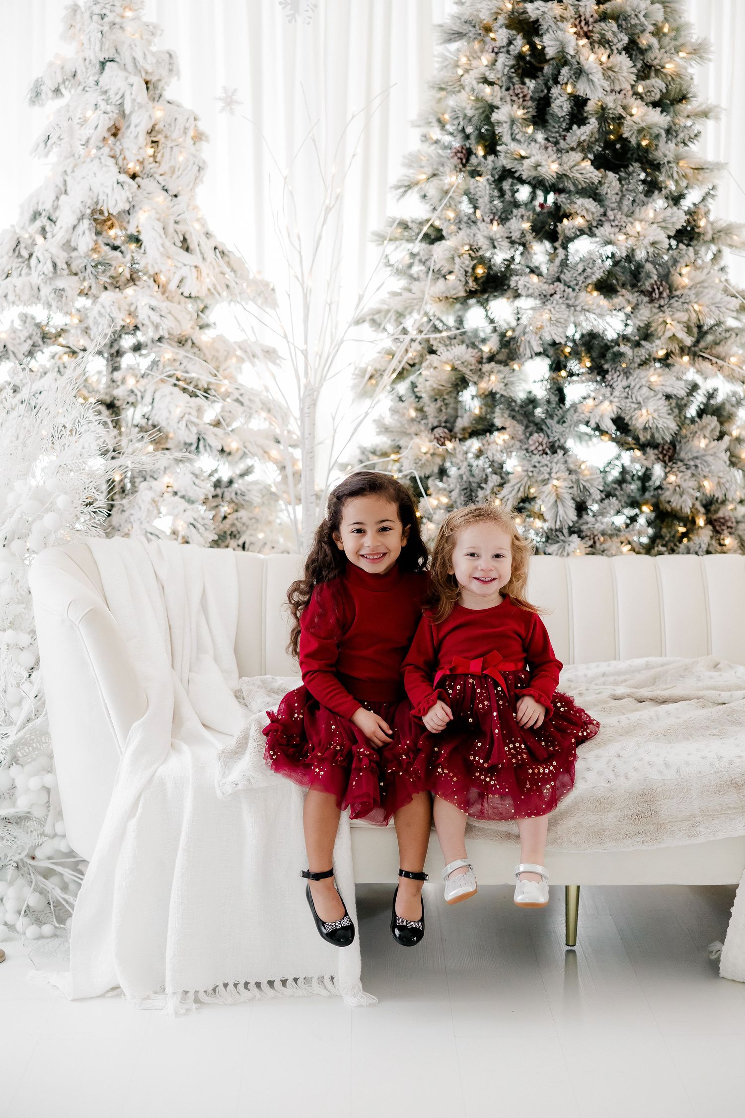 Two young children in red holiday dresses sitting together by white Christmas trees.