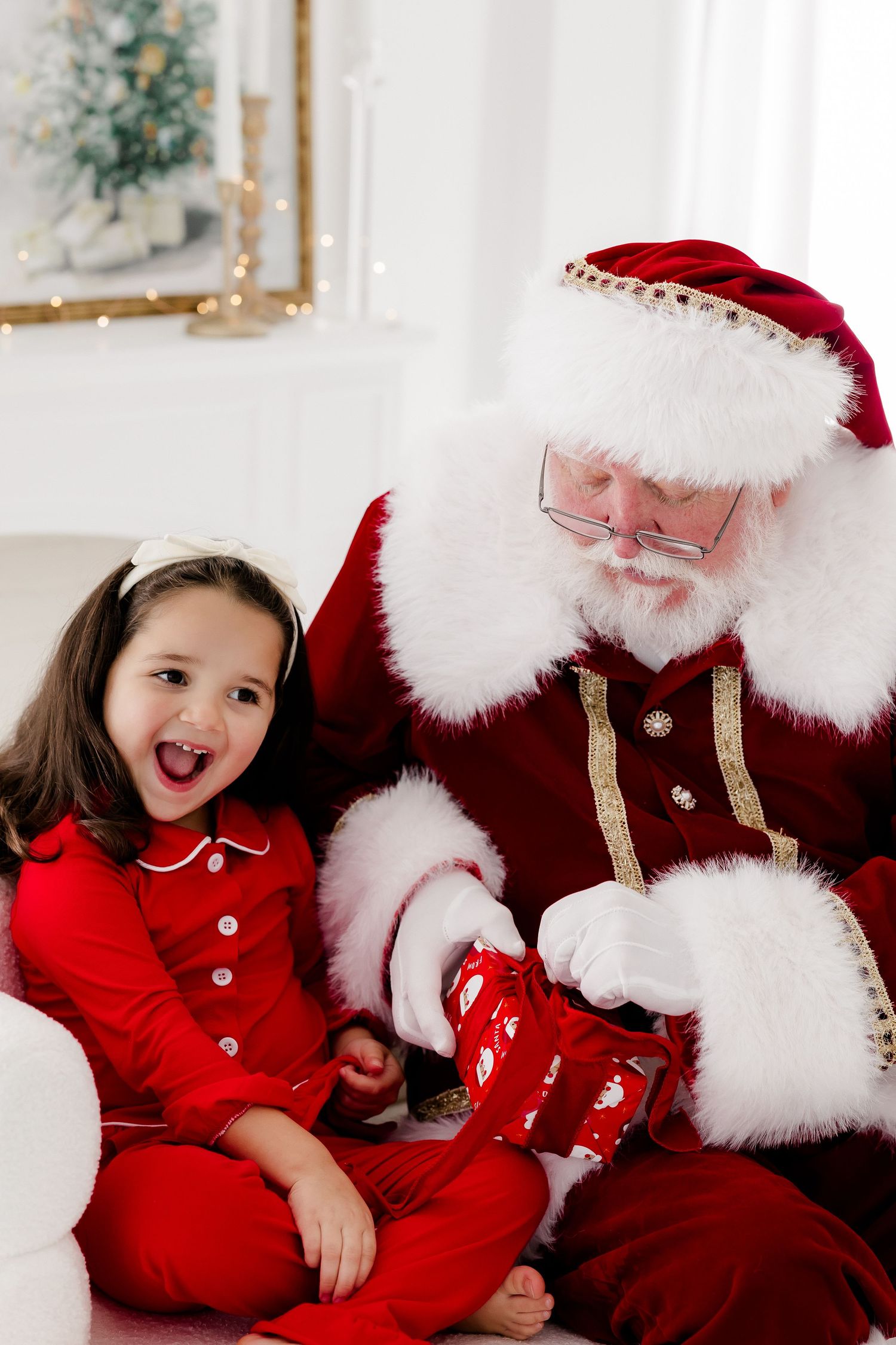 Excited young girl in red pajamas sits with Santa Claus, who opens a Christmas stocking in a festive white room.