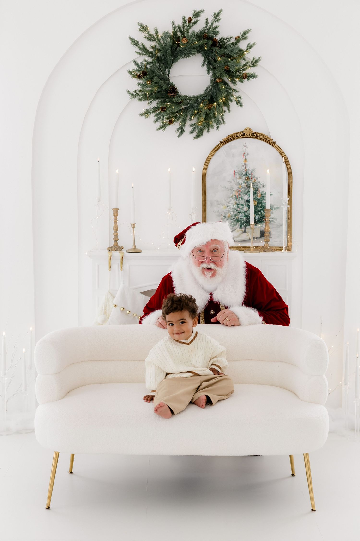 Santa Claus sits on a white sofa with a young child in a festive white room decorated with a Christmas wreath.