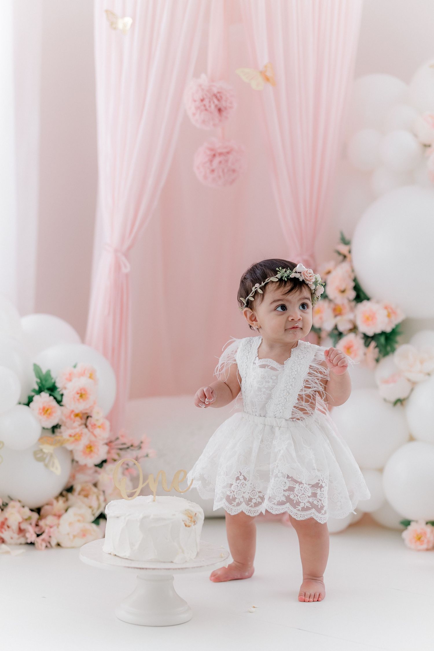 Sweet cake smash photography setup with white ruffled dress, pink floral decorations and soft lighting.