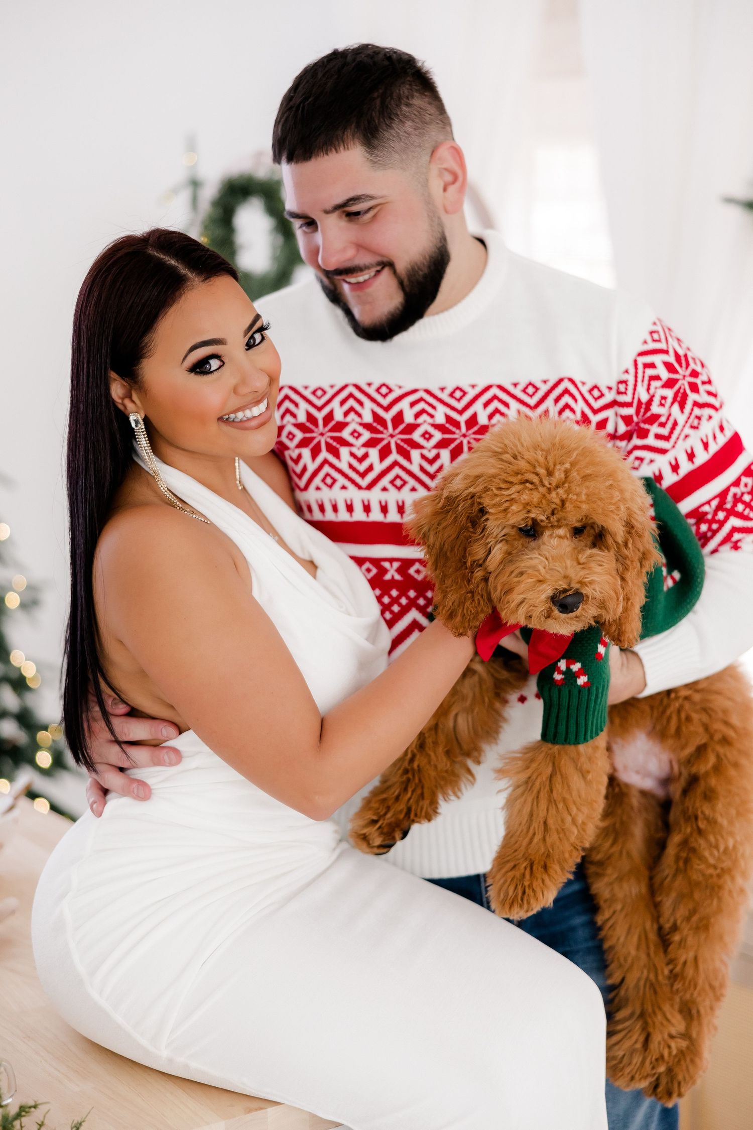 Couple in holiday sweaters cuddling with their goldendoodle dog against white background.