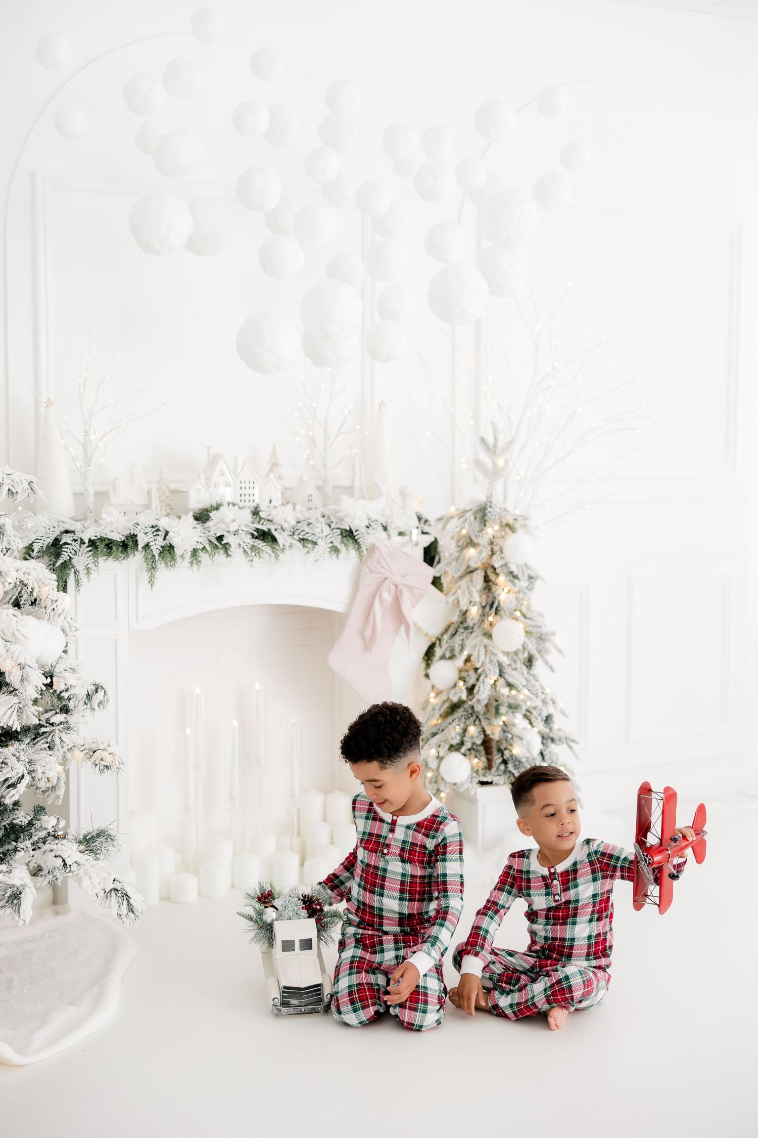 Two children in matching plaid pajamas sitting by festive white fireplace with Christmas decorations.