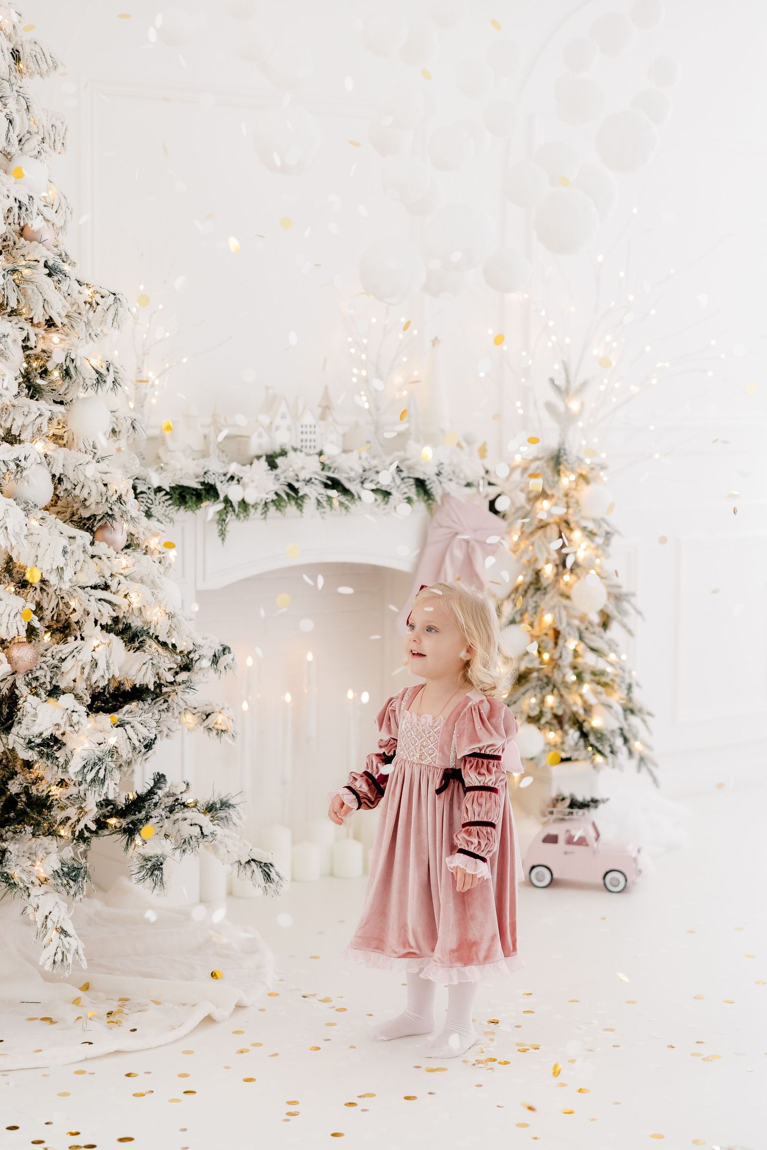 Little girl in pink dress plays in magical winter wonderland setting with flocked Christmas trees.