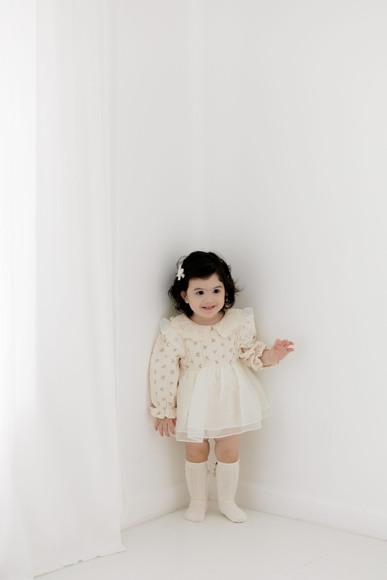 Toddler girl in cream dress and knee socks smiling in white corner studio photo.