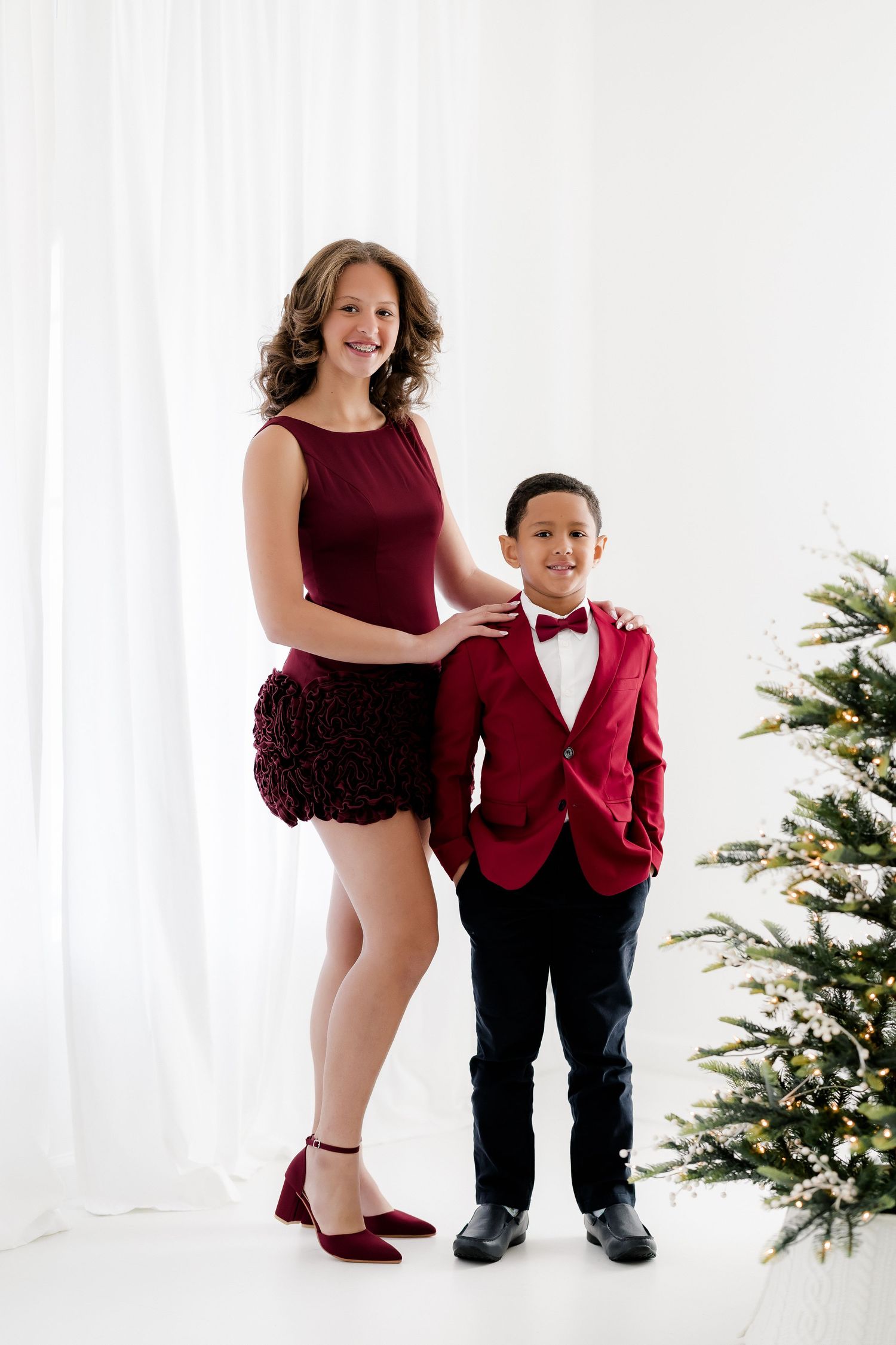 Woman and boy in matching burgundy outfits posing by a Christmas tree in a white studio setting.