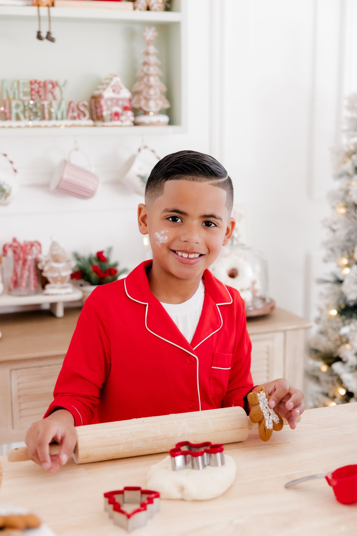 A smiling boy in red Christmas pajamas holds a gingerbread man and rolling pin while baking holiday cookies.