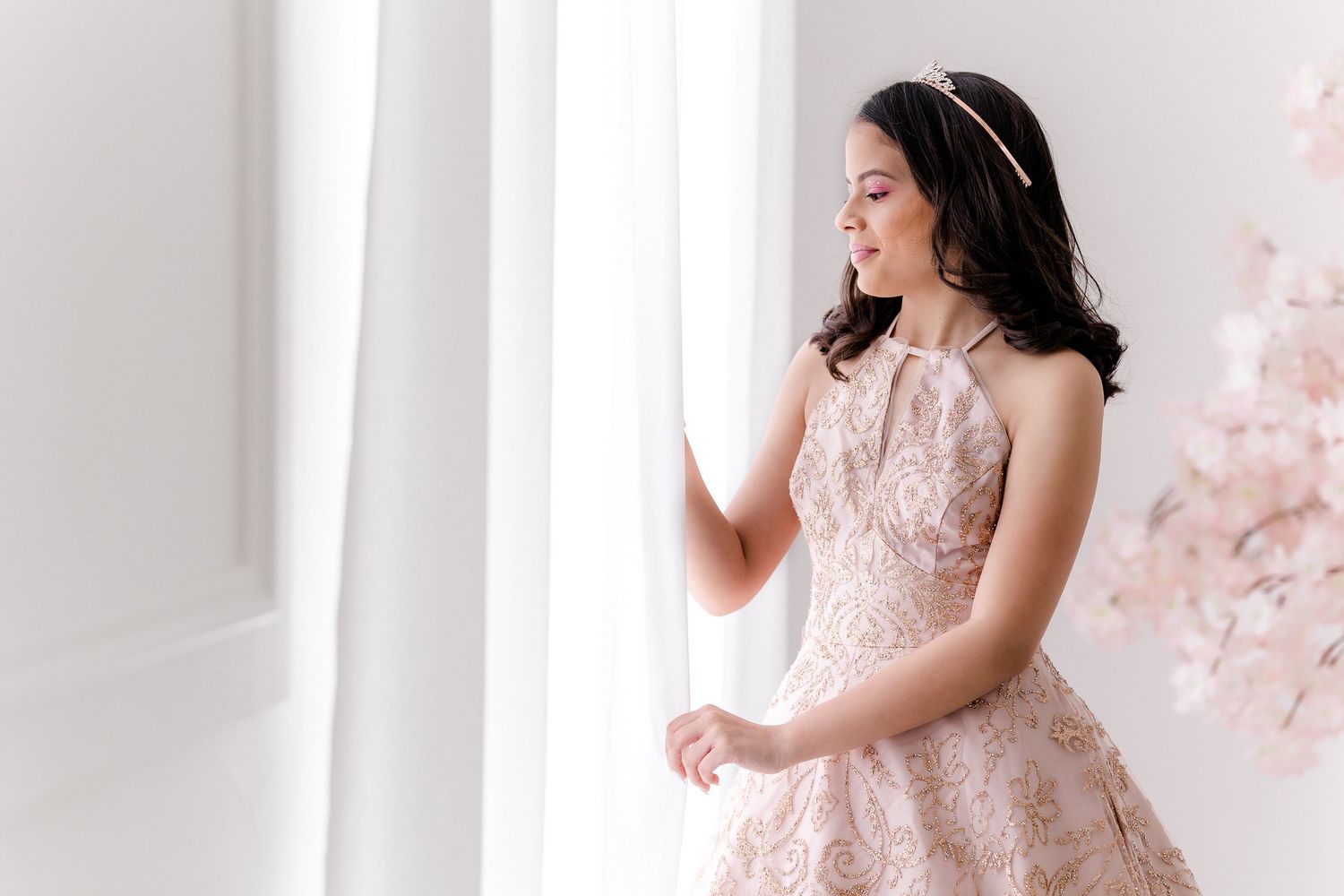 A woman in a romantic pink lace maternity gown poses gracefully by a window with soft natural light.