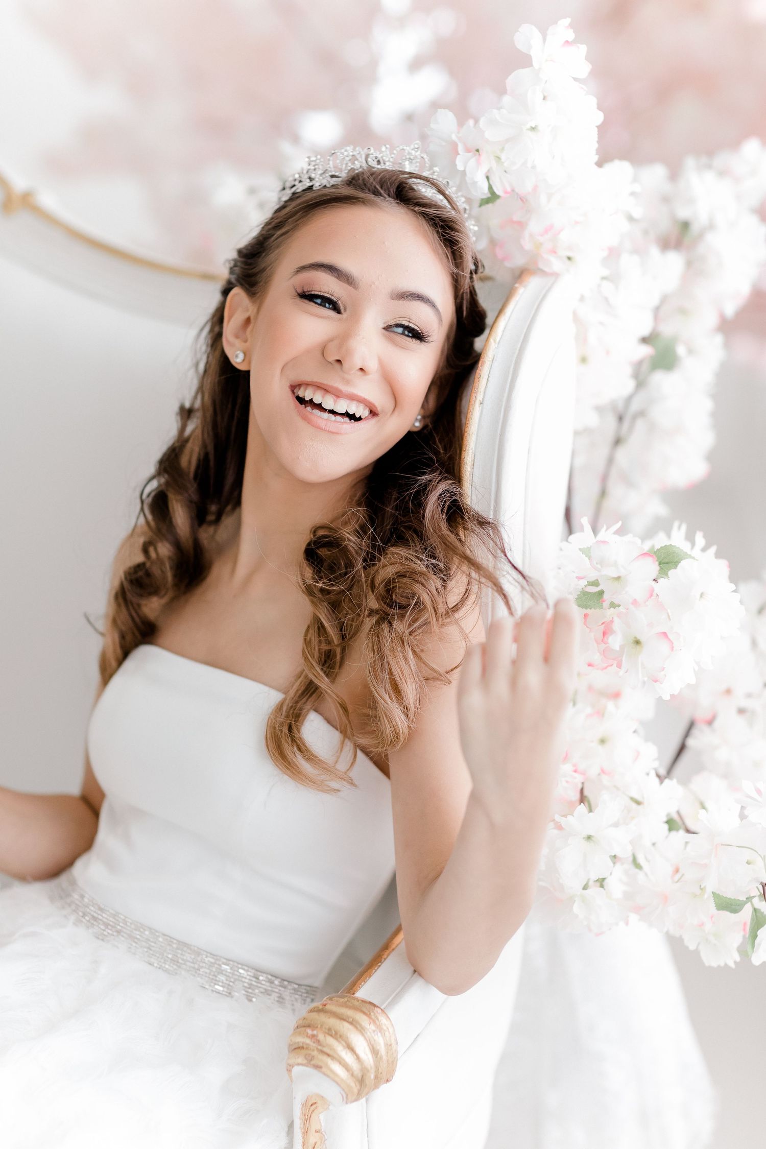 A joyful bride in a white dress and tiara poses with delicate pink cherry blossoms against a light background.