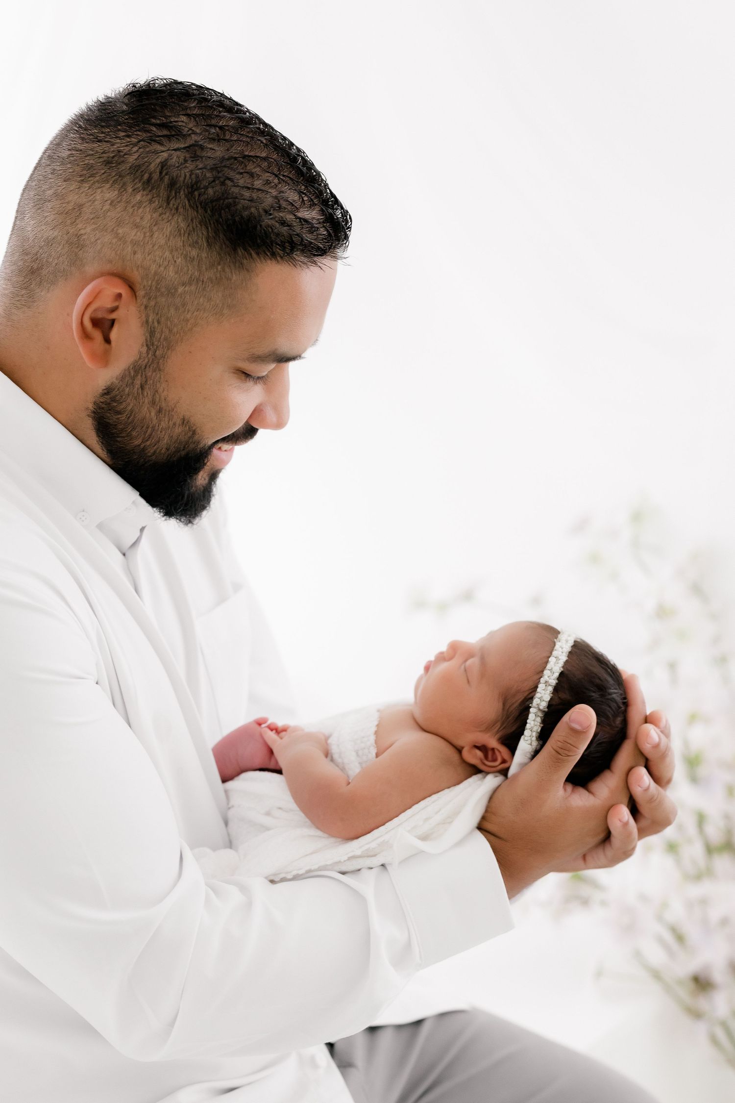 A tender moment between a parent in white holding a sleeping newborn baby against a bright background.