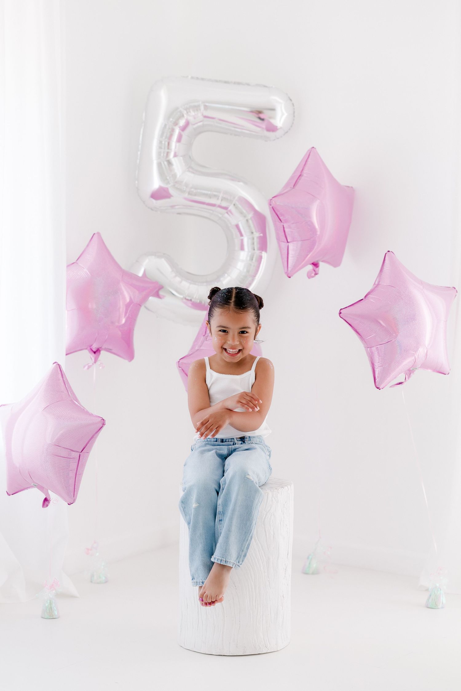 A baby in light denim sits among pink star balloons and white decor in a minimalist photo studio setting.