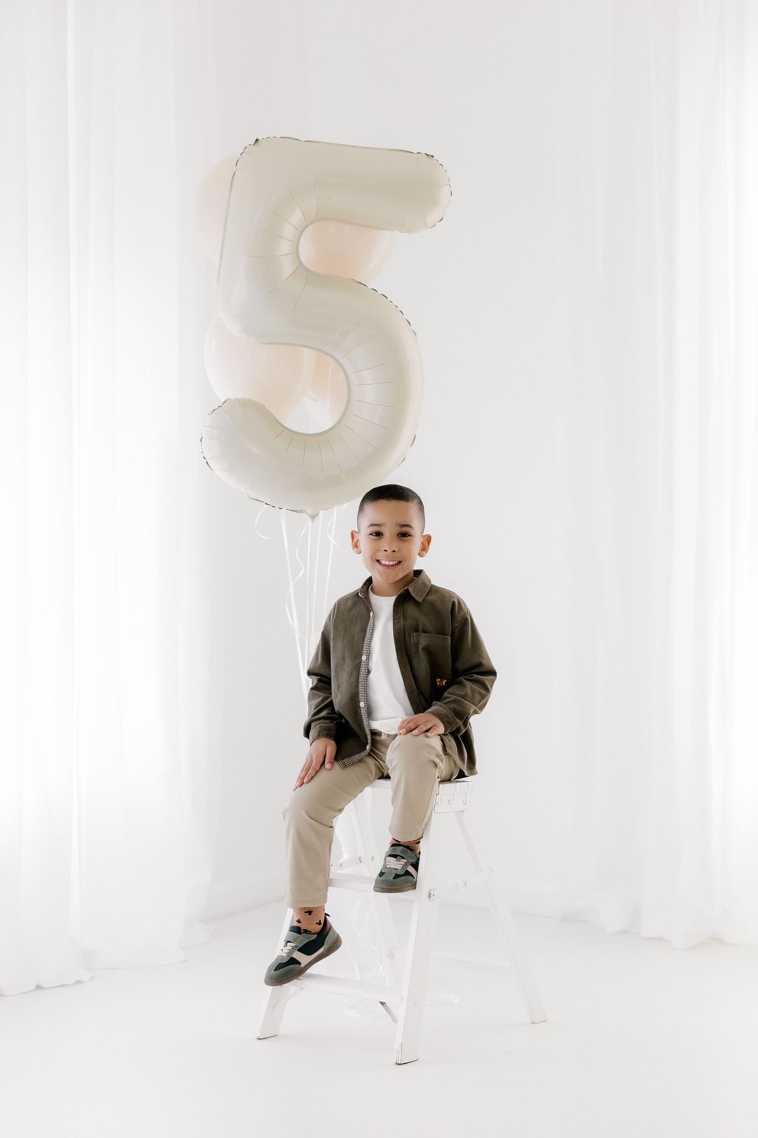 Child sitting on white chair holding large number 5 balloon in minimalist studio setting.