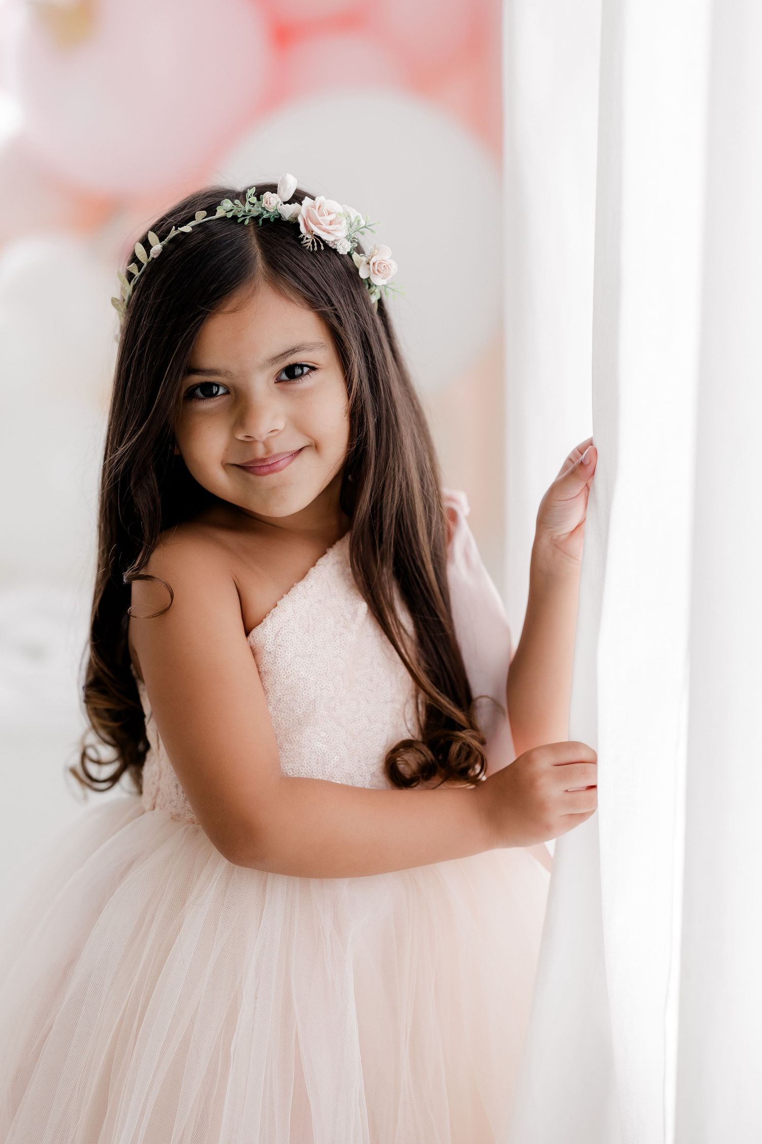 Young girl in white lace dress with tulle skirt poses gracefully between white pillars in soft lighting.