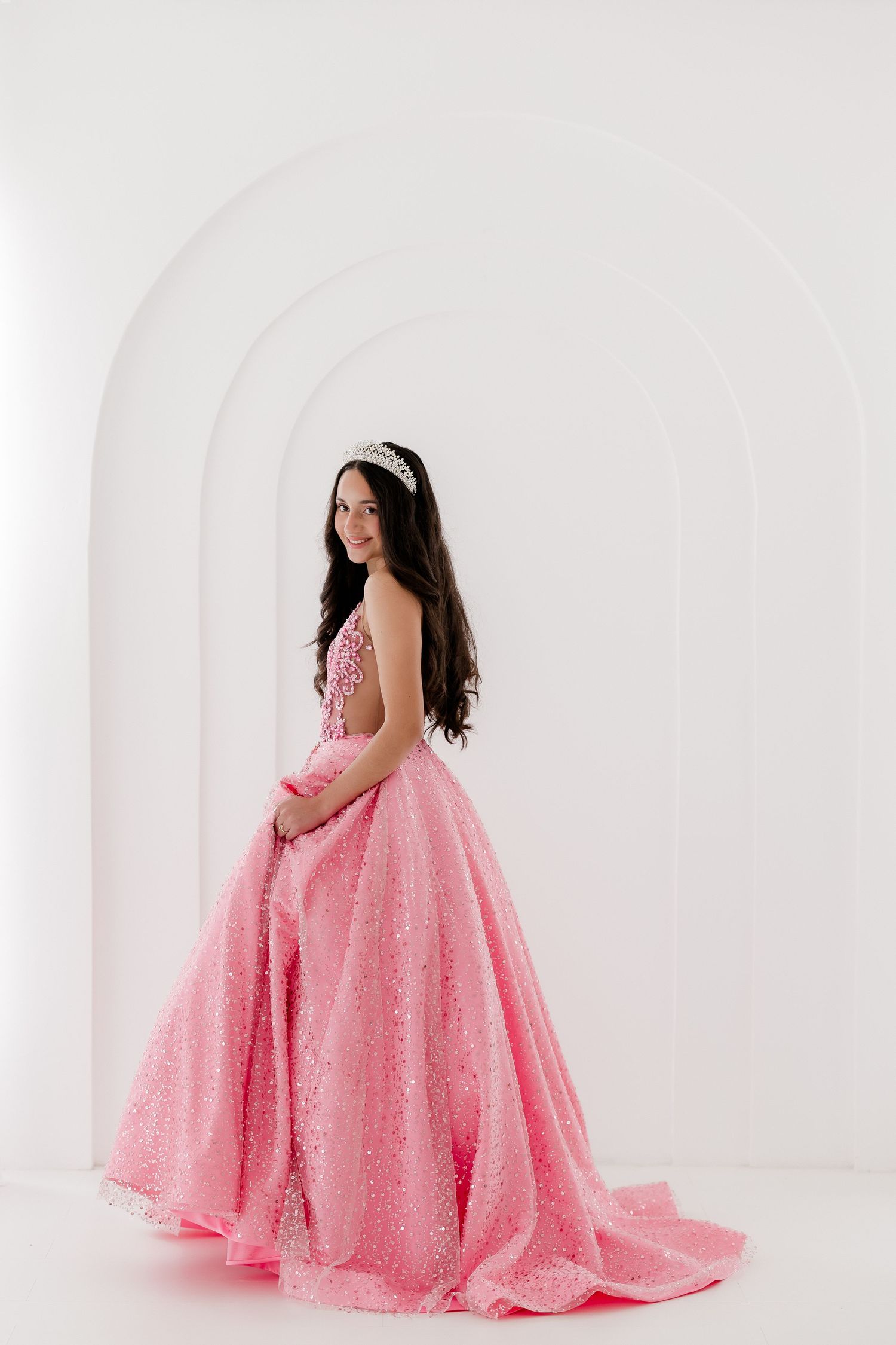 Expectant mother in flowing pink glitter gown poses gracefully against white backdrop in maternity photoshoot.