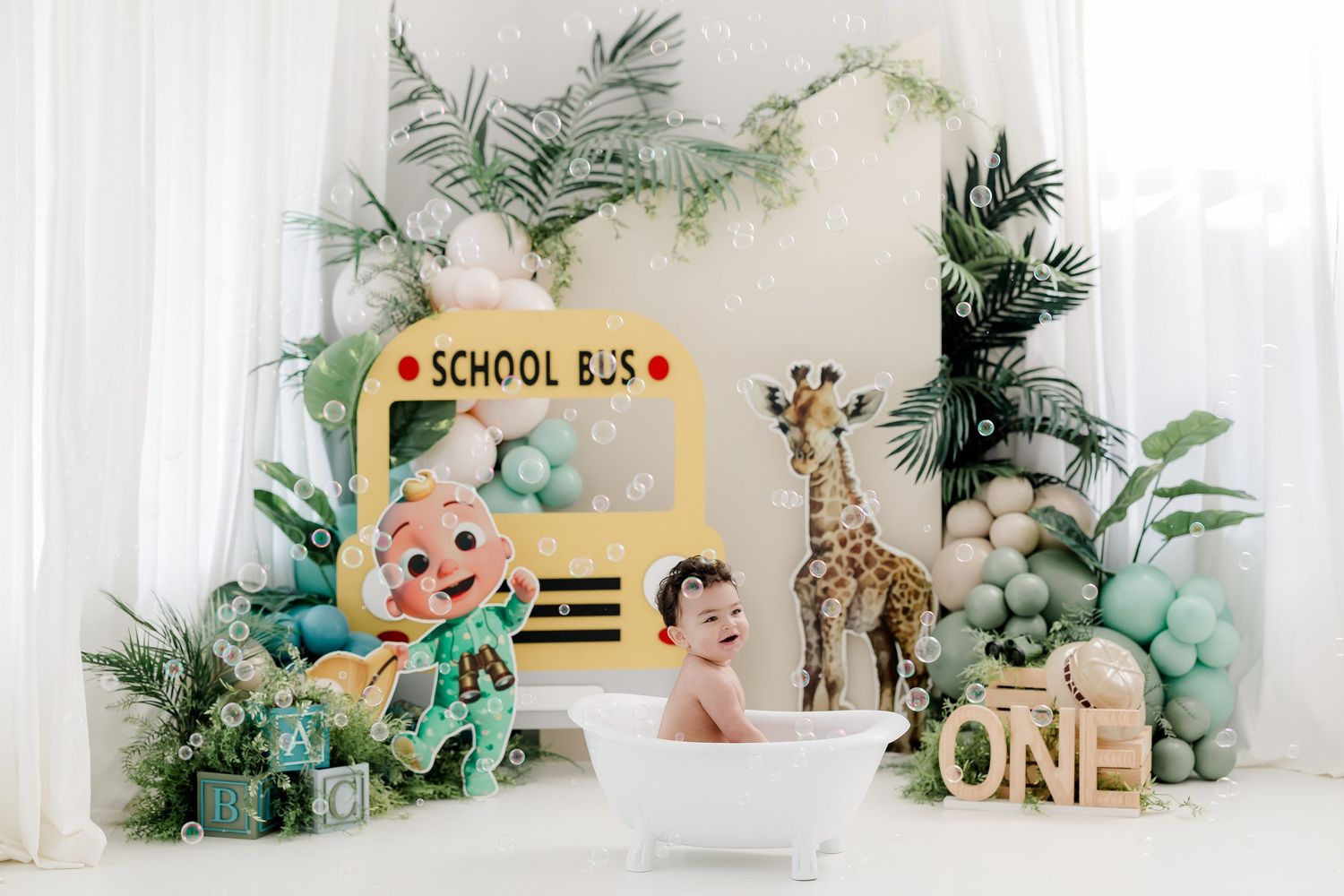 Safari-themed first birthday party decorations with green foliage, balloons, and jungle animal cutouts against a white backdrop.
