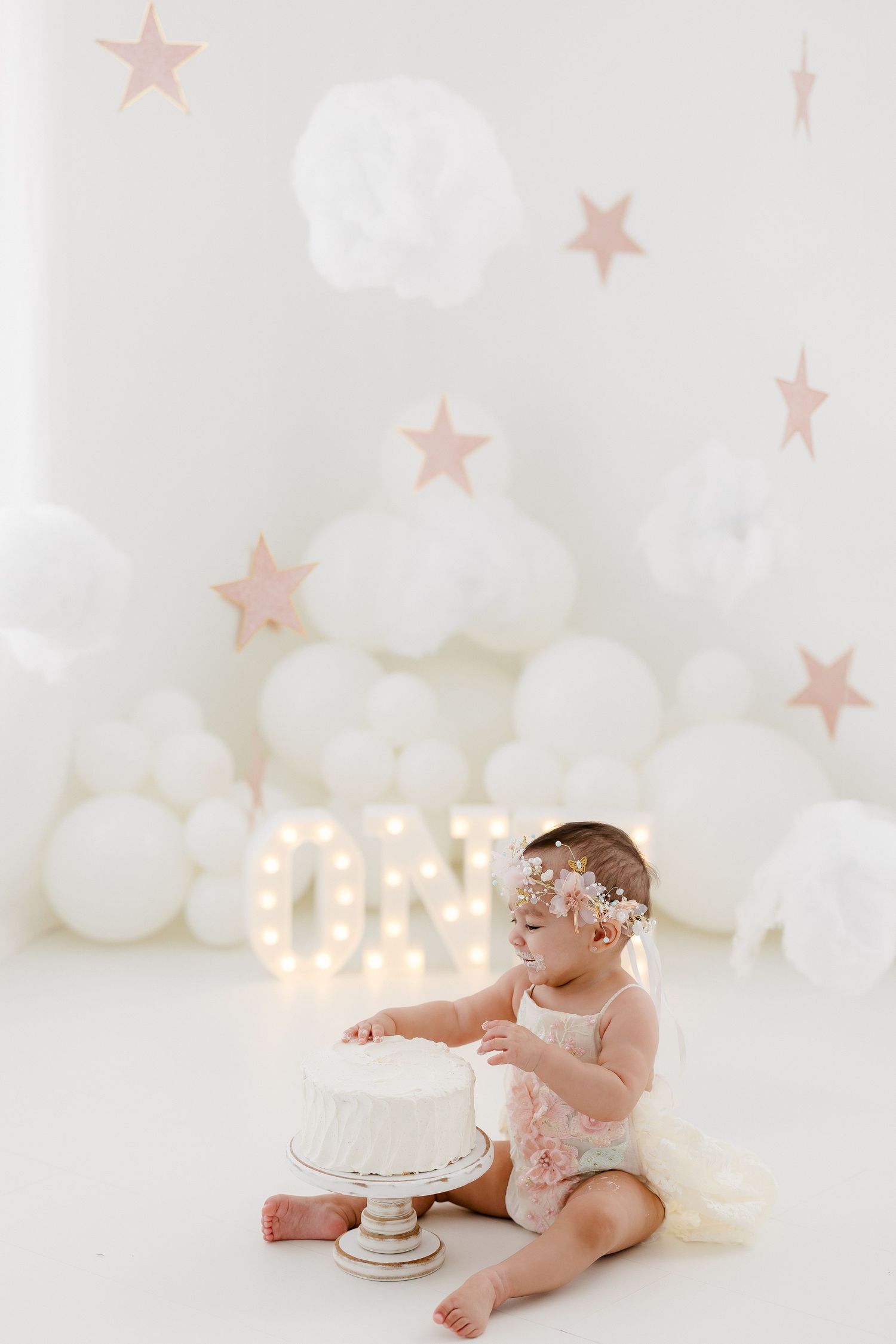 A cake smash photo session setup with white balloons, stars, and glowing letters against a light backdrop.