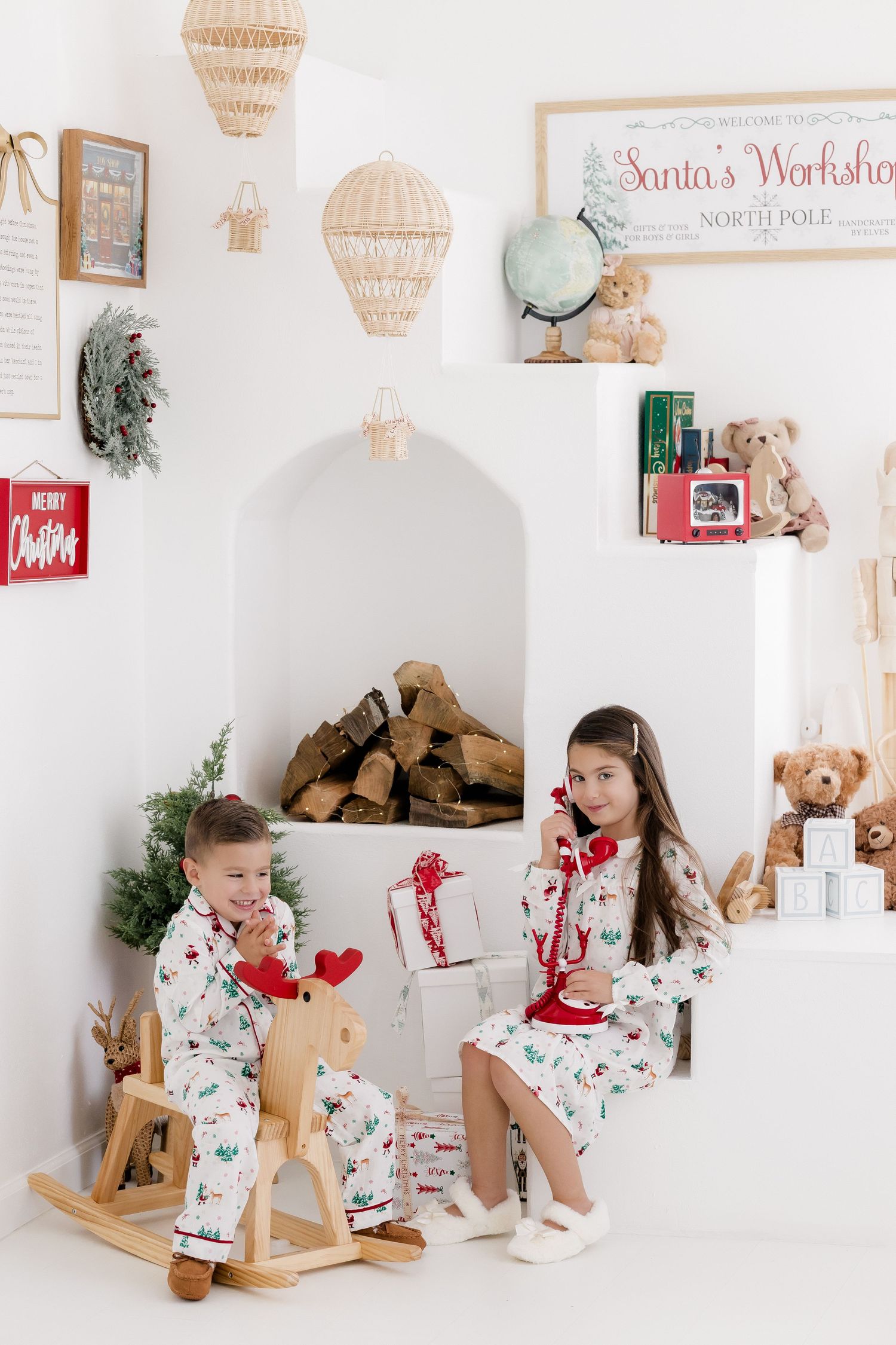 Two children in matching Christmas pajamas sit near a festive Santa's Workshop display with toys and gifts.