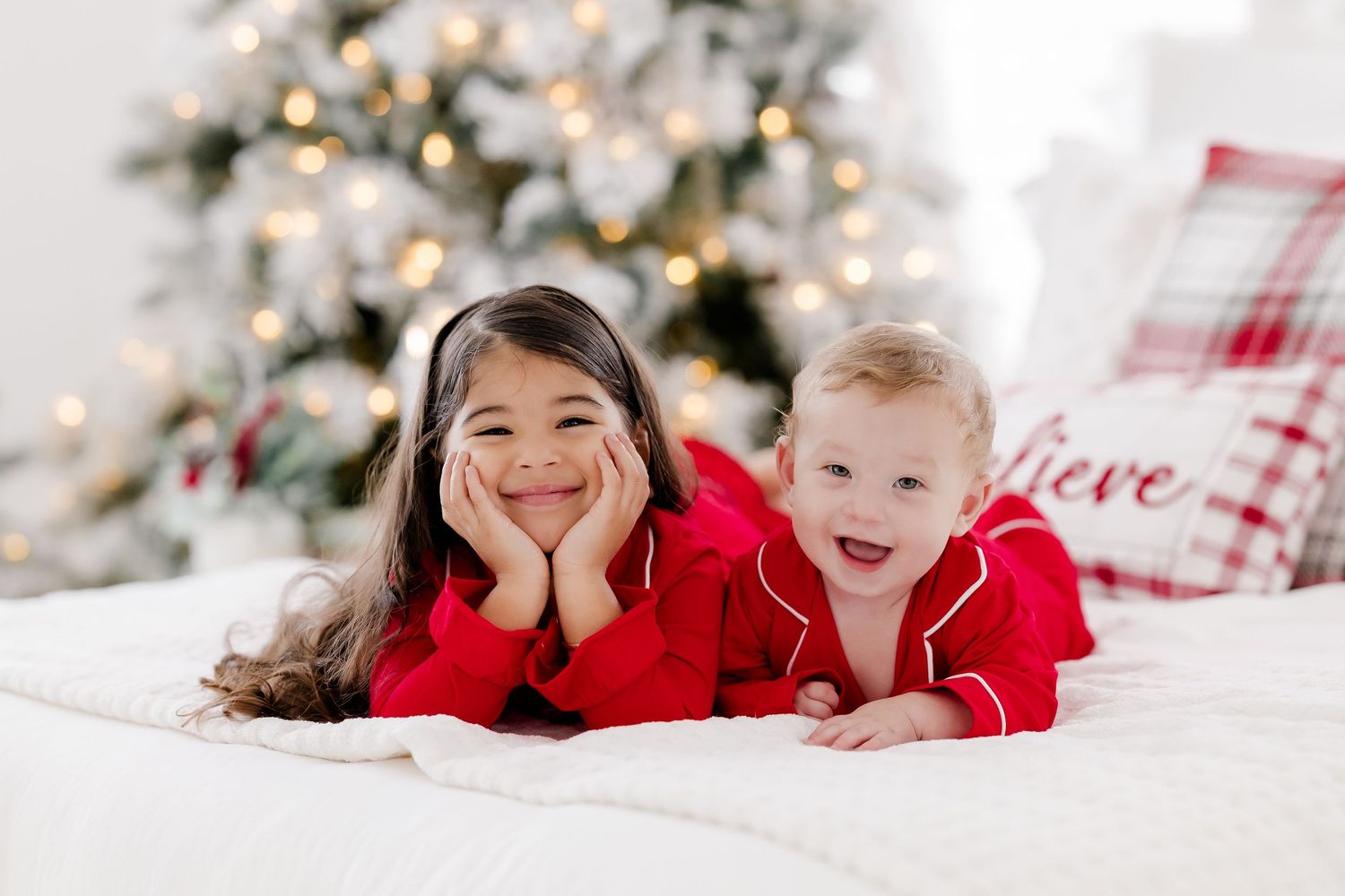 Two children in red Christmas pajamas lying on a bed smiling, with a decorated Christmas tree in the background.