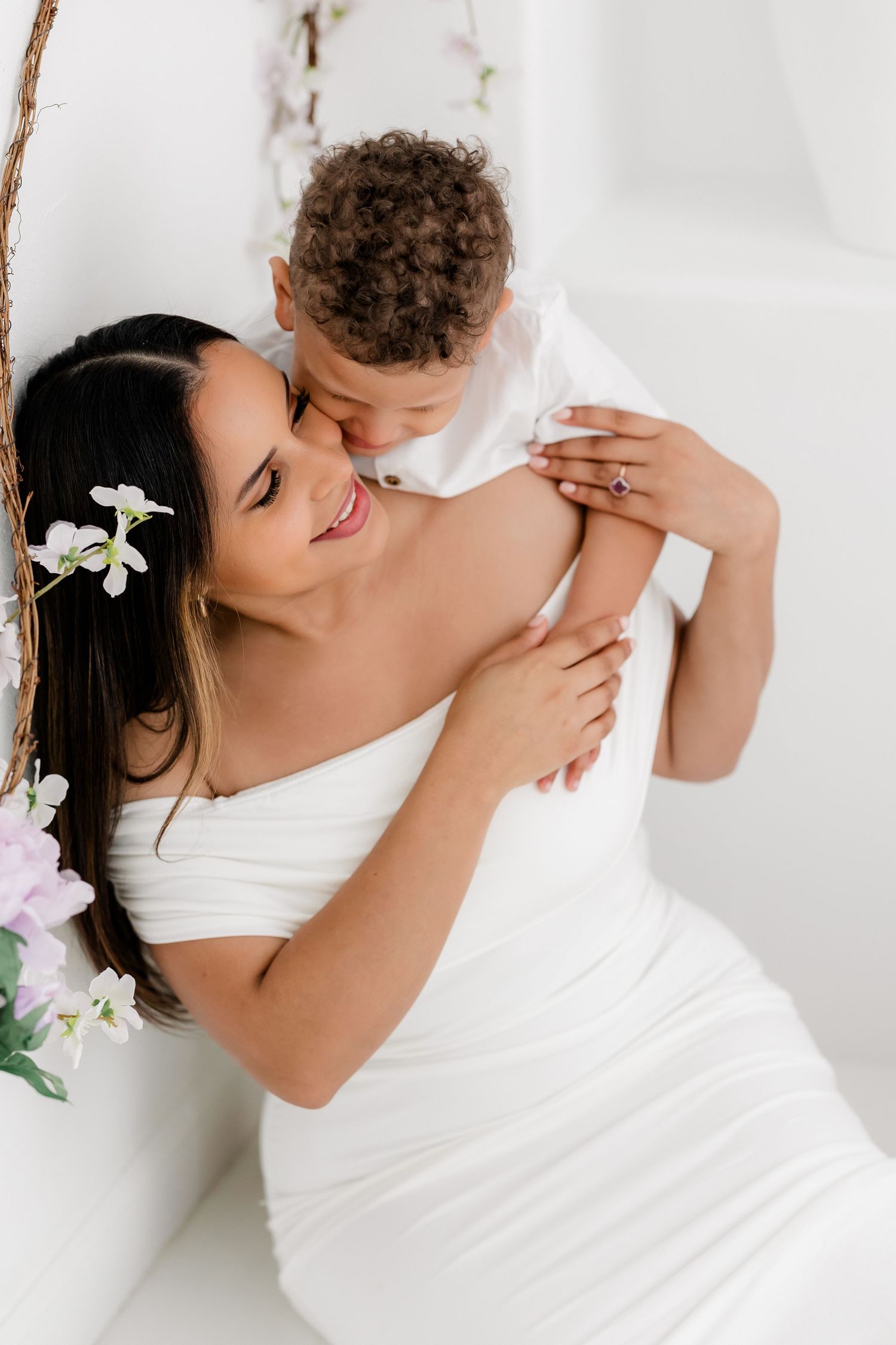 Tender embrace between couple in white against light background with delicate white flowers in her dark hair.