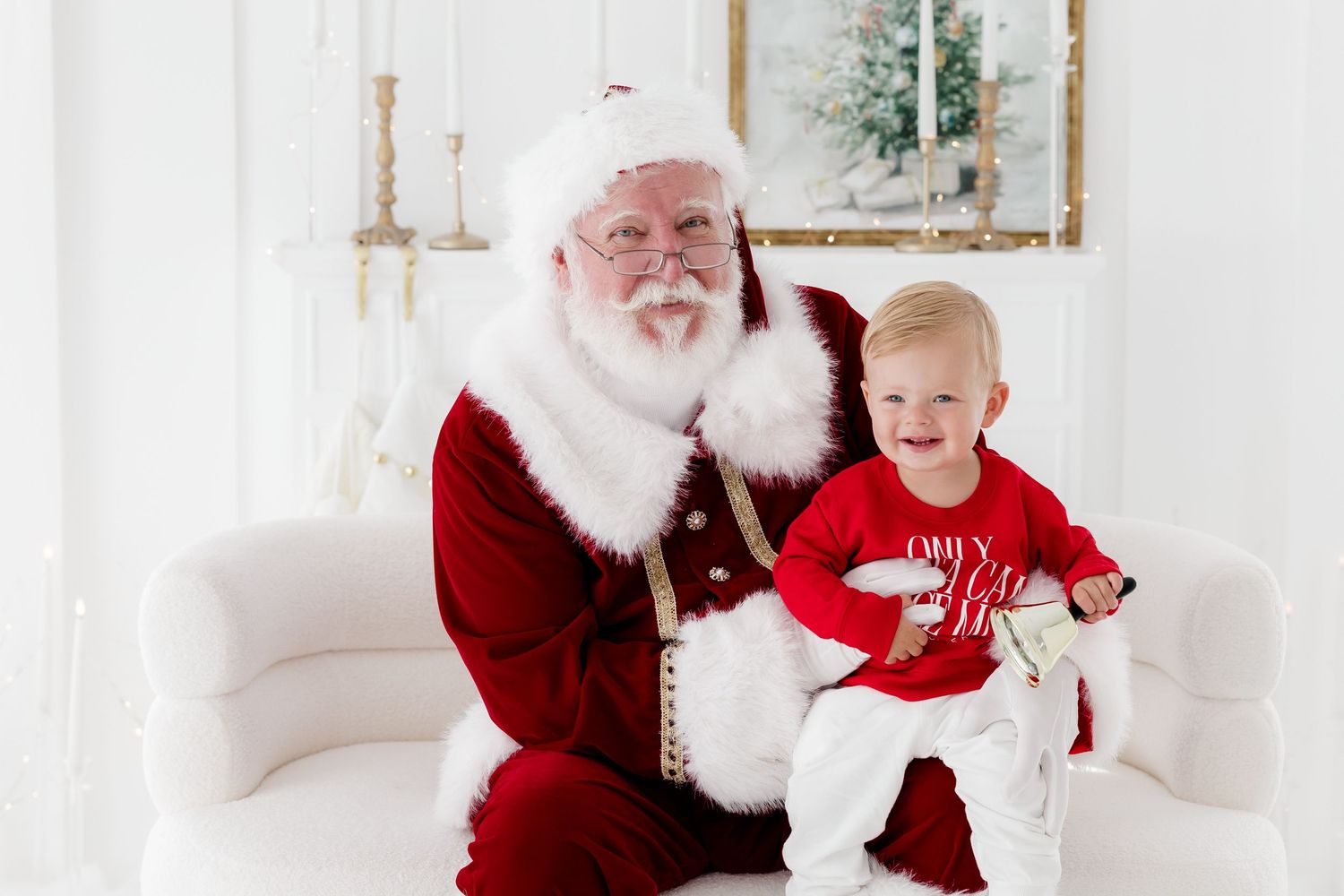 A cheerful toddler sits on Santa Claus's lap in a bright white studio during a festive Christmas photo session.