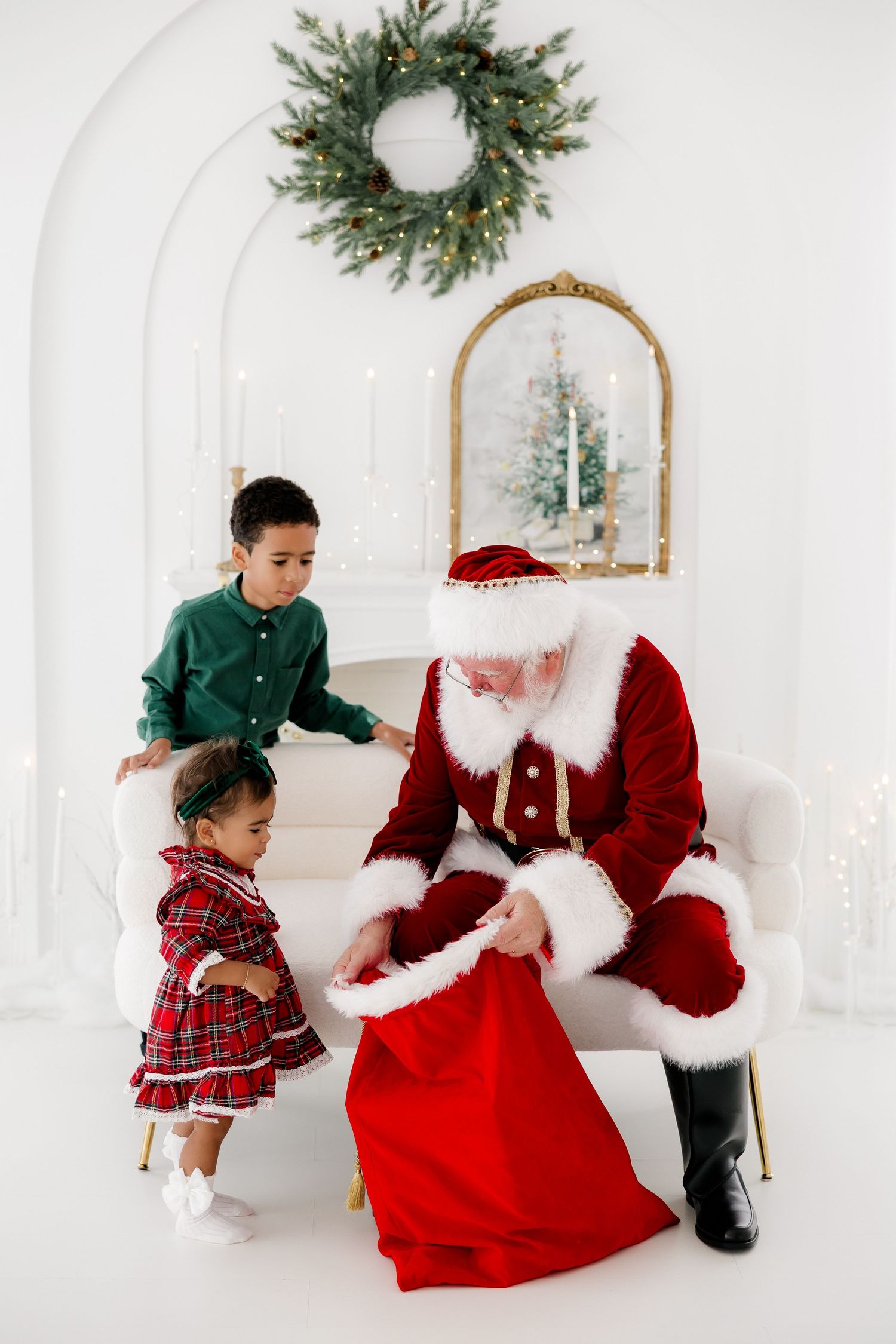 Santa Claus sits on a white sofa with two young children, reaching into his red gift bag in a festive white decorated room.